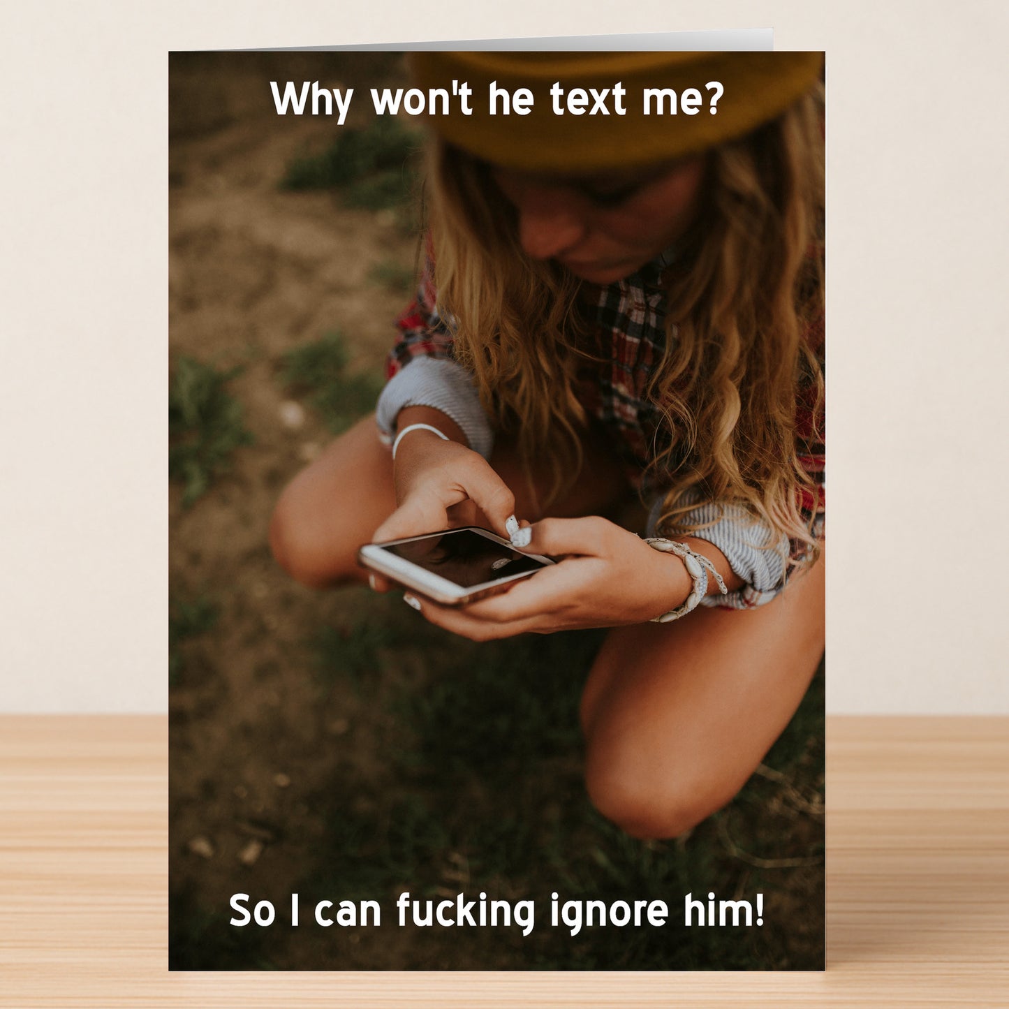The Ignore Funny Greeting Card by Twisted Gifts features a woman outdoors checking her phone with the words, "Why won't he text me?" above and, "So I can fucking ignore him!" below. A witty, high-quality card for a good laugh.