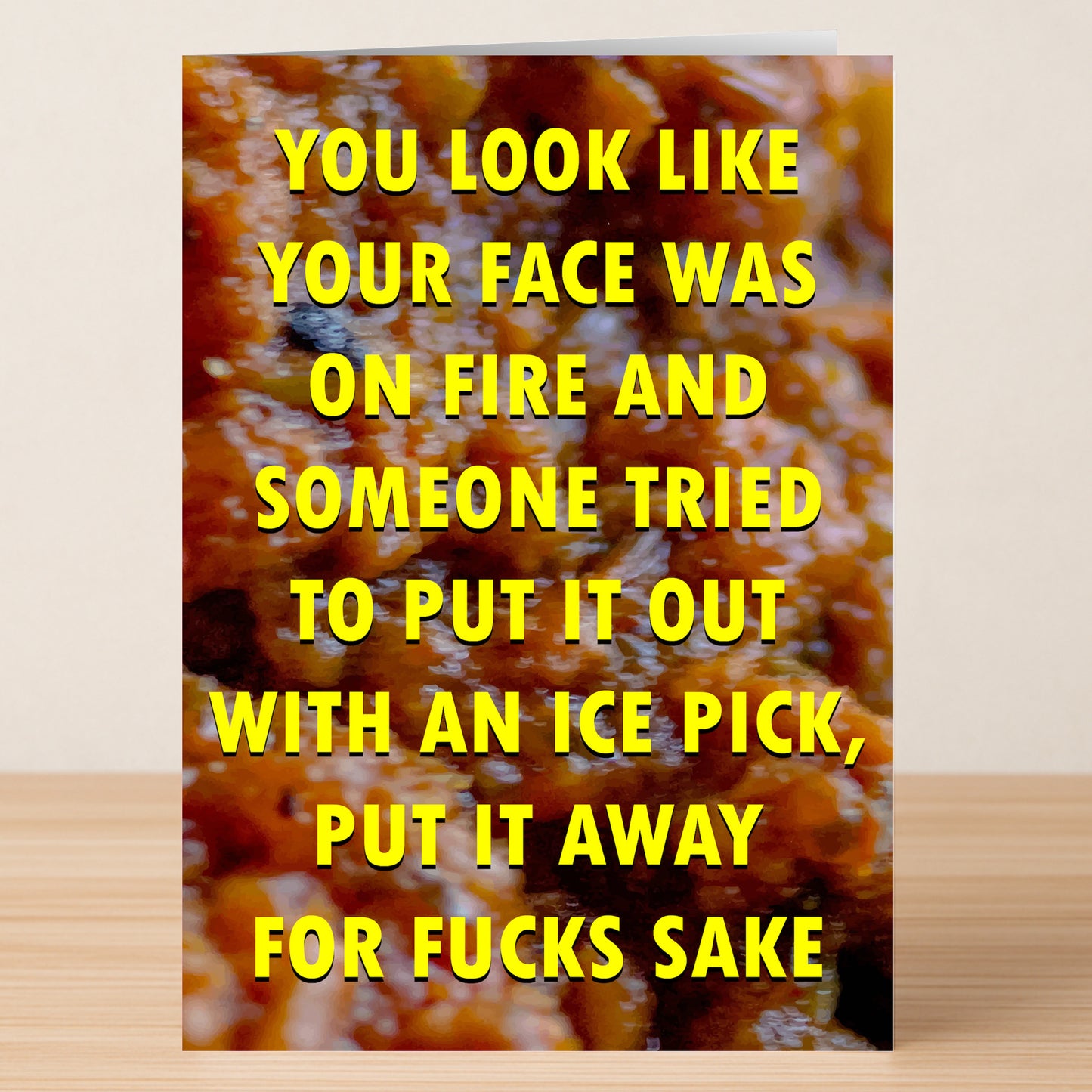 The Ice pick Insulting Greeting Card by Twisted Gifts features a bold yellow message: "YOU LOOK LIKE YOUR FACE WAS ON FIRE AND SOMEONE TRIED TO PUT IT OUT WITH AN ICE PICK, PUT IT AWAY FOR FUCKS SAKE," and sits on a wooden surface.
