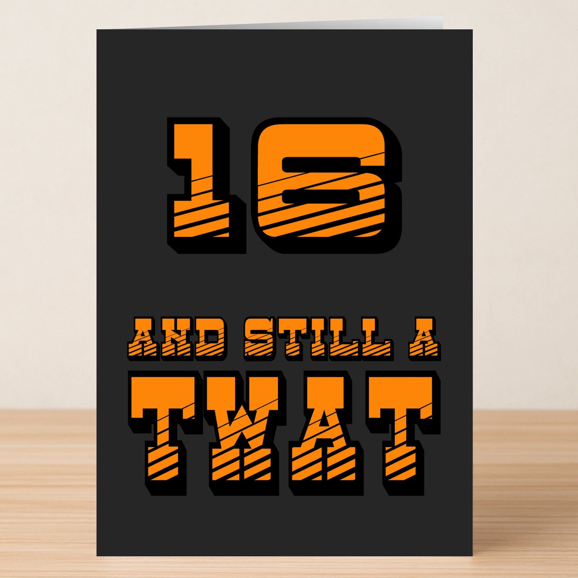 A funny "16 - Still A Twat" birthday card from Twisted Gifts with bold orange text on black, displayed on a wooden surface—perfect for anyone who enjoys a playful, cheeky card.