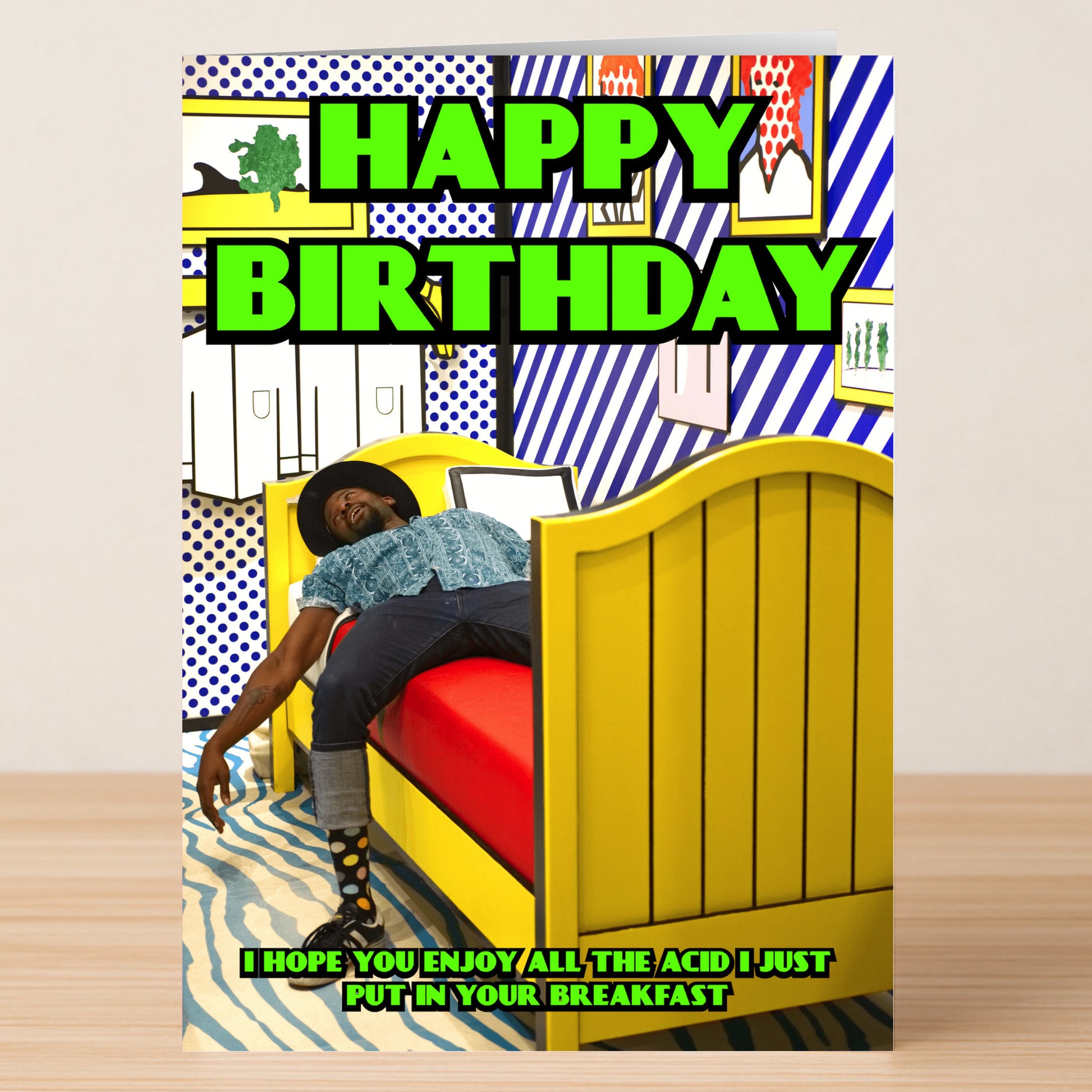The Breakfast Funny Birthday Card by Twisted Gifts shows a person lounging on a bright yellow bed in a vibrant room, with “HAPPY BIRTHDAY” and a witty joke about adding acid to breakfast for extra humor.