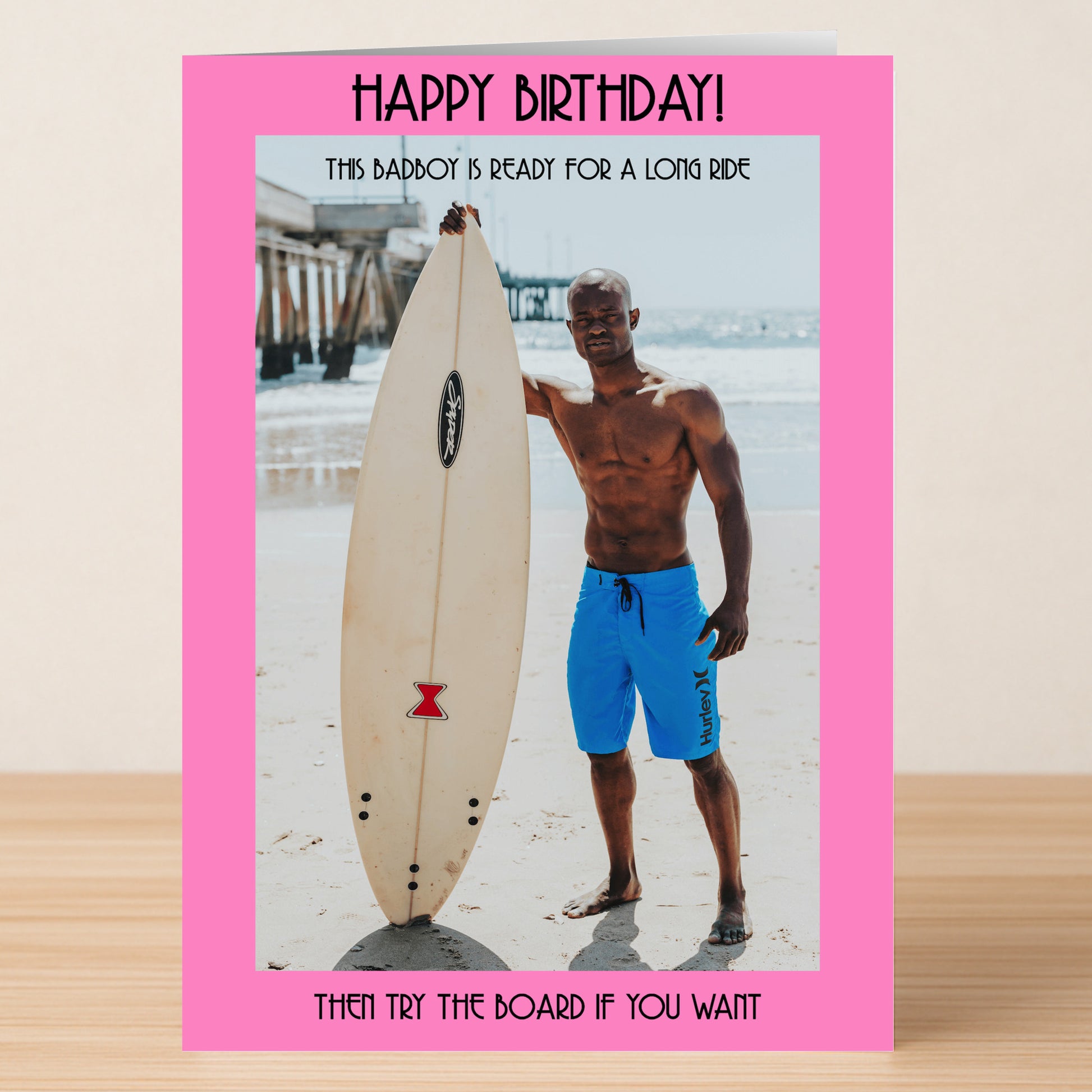 The Ready For A Ride Funny Birthday Card by Twisted Gifts shows a man in blue shorts on a beach beside a surfboard with a pier behind him, framed by a pink border and witty birthday message.