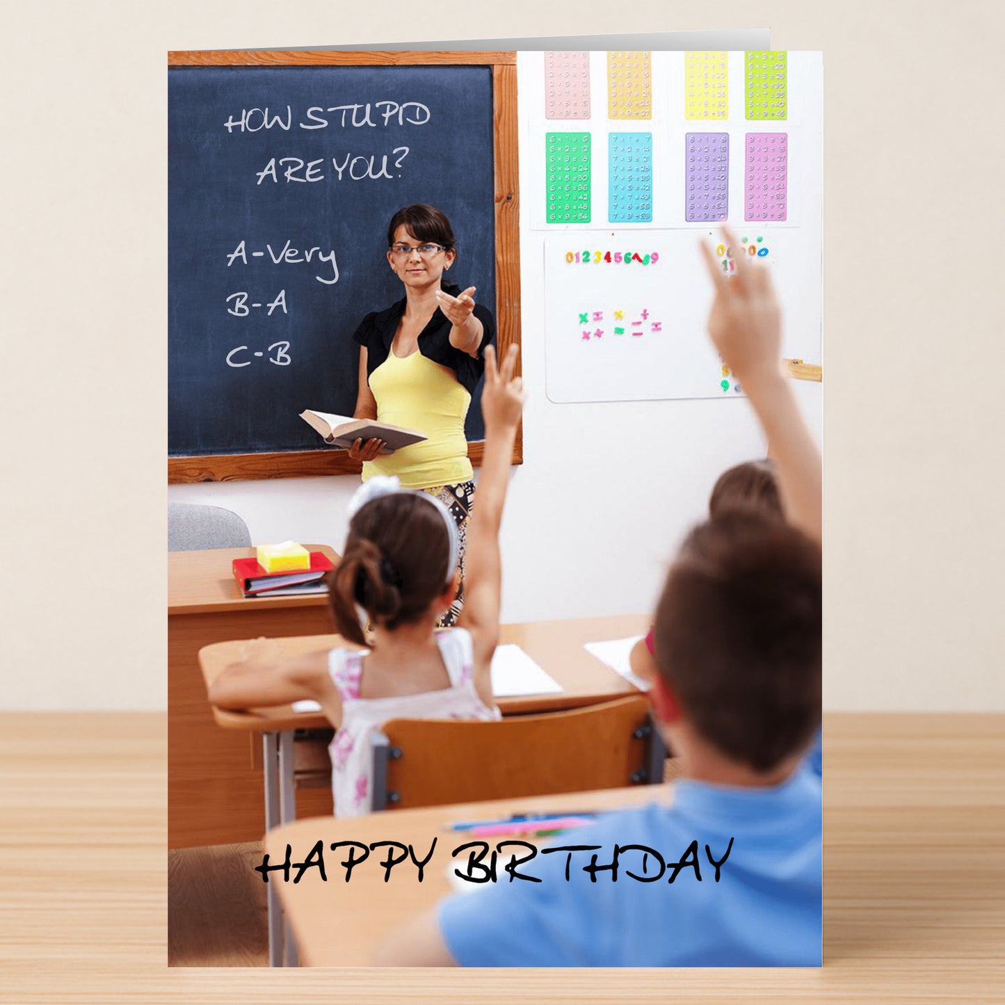 A teacher points to students with raised hands in front of a blackboard that says "HOW STUPID ARE YOU?" above "HAPPY BIRTHDAY"—the How Stupid Insulting Birthday Card by Twisted Gifts is perfect for fans of cheeky, funny birthday cards.