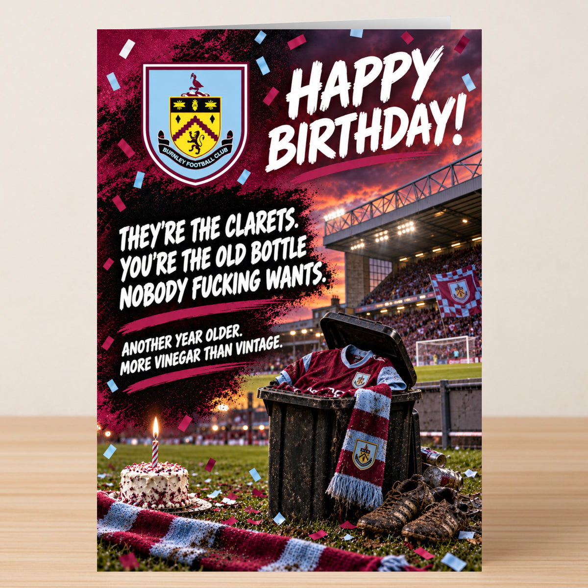A Burnley – Funny Birthday Card featuring the Burnley FC crest, a trash can filled with a jersey and scarf, a small cake on a grass pitch, and the text “Happy Birthday! They’re the clarets.”