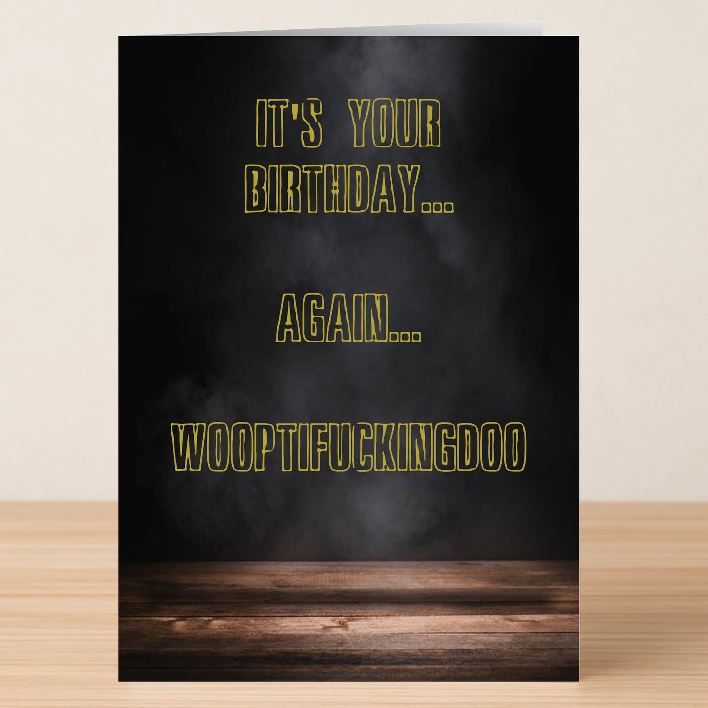 The Woopti Insulting Birthday Card by Twisted Gifts features a black smoky background, wooden surface, and bold yellow-outlined text: "IT'S YOUR BIRTHDAY... AGAIN... WOOPTIFUCKINGDOO"—a funny card that says it all!.