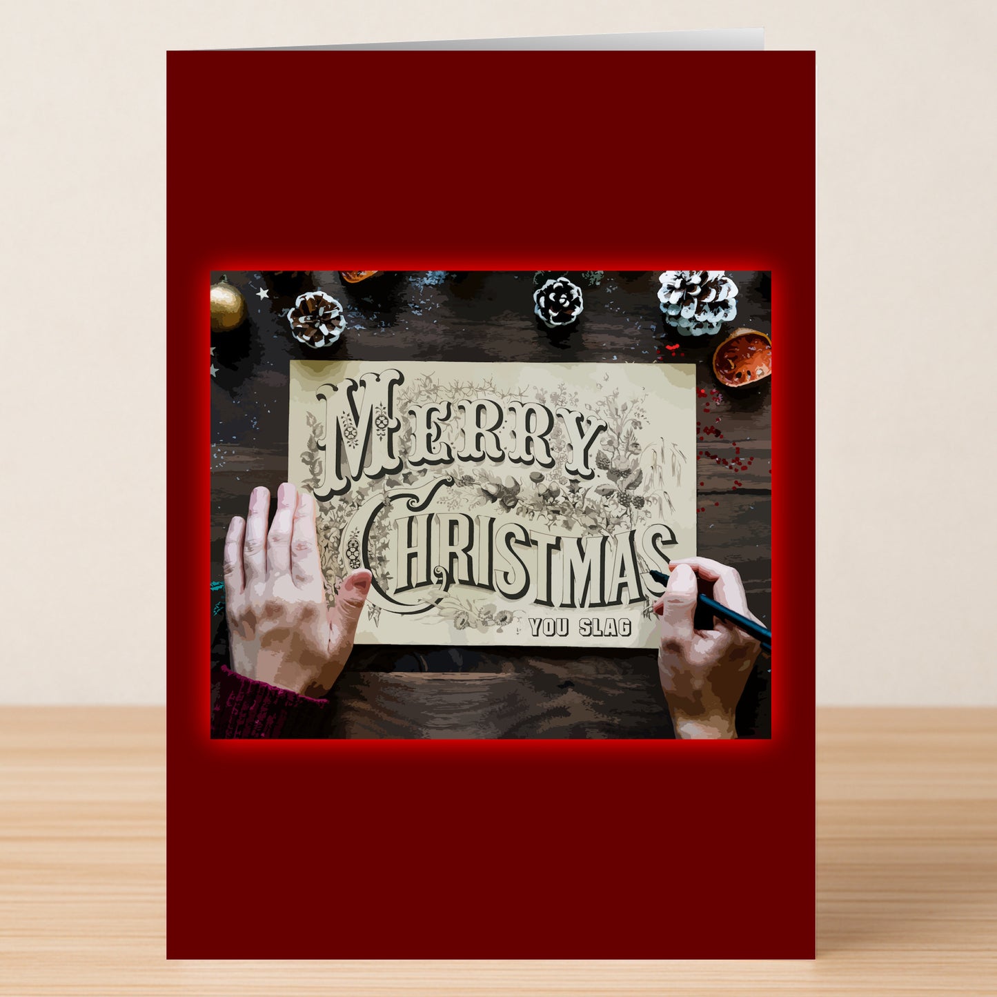 Someone writes on the You Slag Rude Christmas Card by Twisted Gifts, featuring "Merry Christmas You Slag" and festive designs on a dark wooden table—making for a bold and unique holiday card.