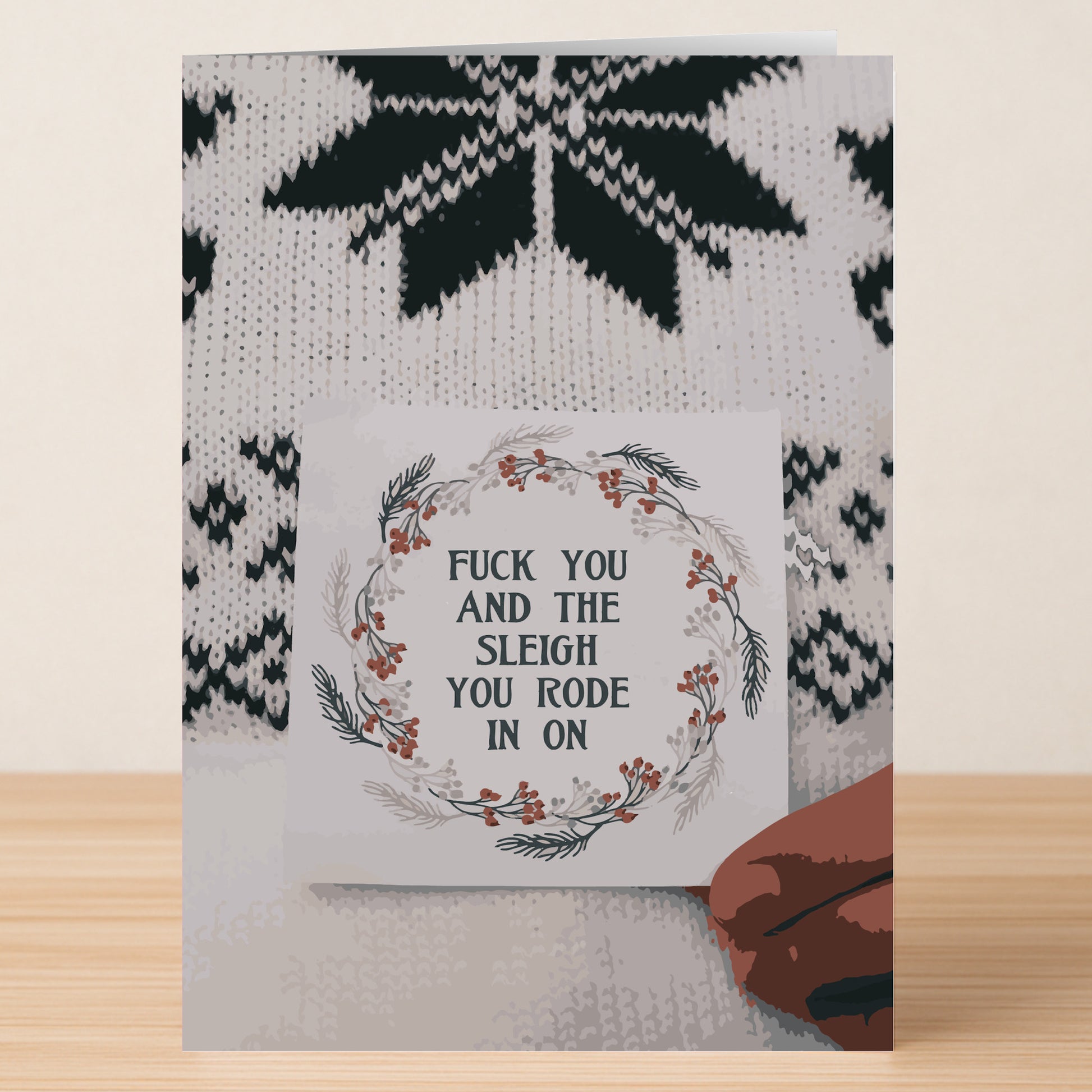 The Twisted Gifts Sleigh Rude Christmas Card rests on a wooden table, featuring the message "FUCK YOU AND THE SLEIGH YOU RODE IN ON" with festive foliage and a knit snowflake-patterned background for cheeky holiday flair.