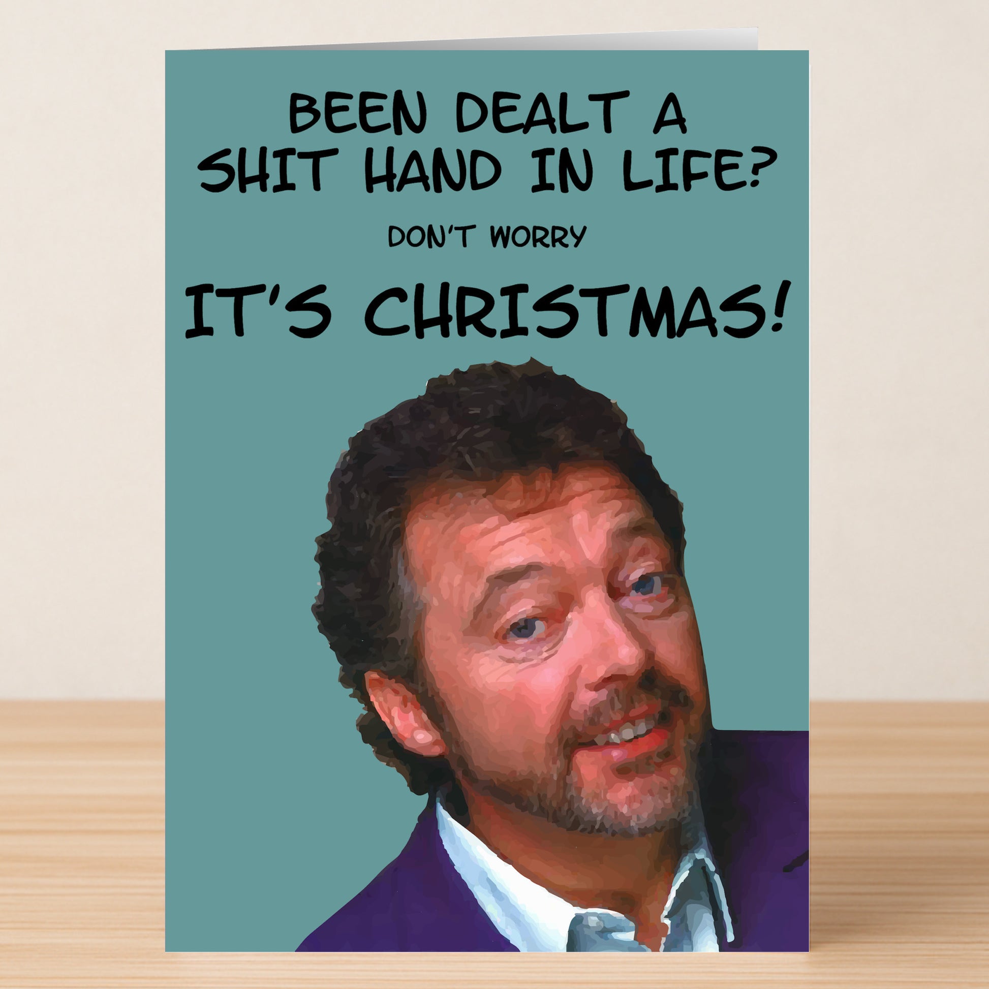 Don't Worry Funny Christmas Card by Twisted Gifts features a man's smiling portrait and the message "Been dealt a shit hand in life? Don't worry IT'S CHRISTMAS!" on a teal background—add humor to your holiday with this unique card.