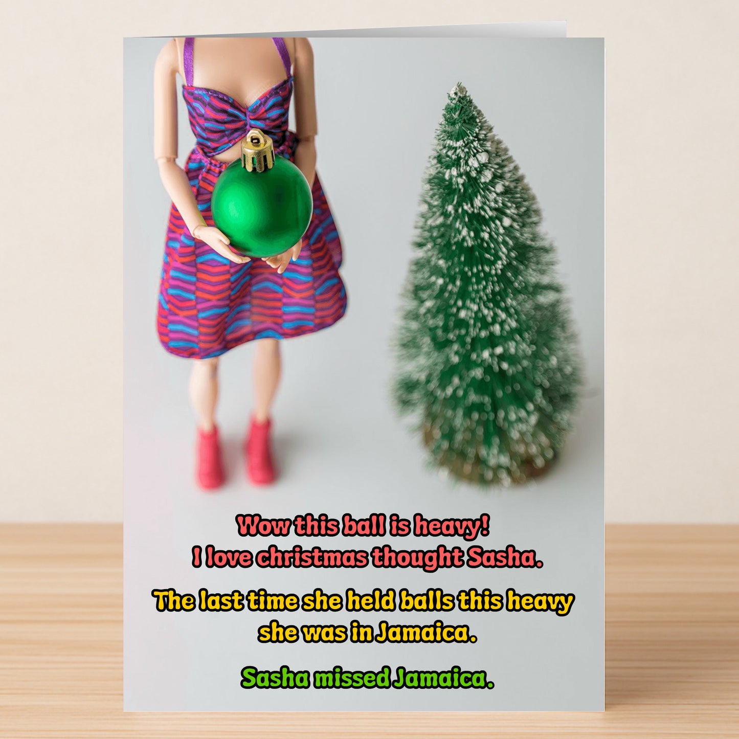 A doll in a bright dress with a big green ornament stands by a small fake Christmas tree on wood, with holiday humor text overlaid—an ideal Jamaica Rude Christmas Card from Twisted Gifts.