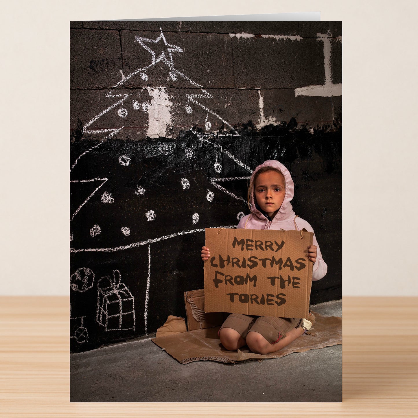 A child sits on cardboard before a chalk Christmas tree, holding a "Merry Christmas from the Tories" sign—it's the Tories Funny Christmas Card by Twisted Gifts, perfect for sending a humorous festive message.