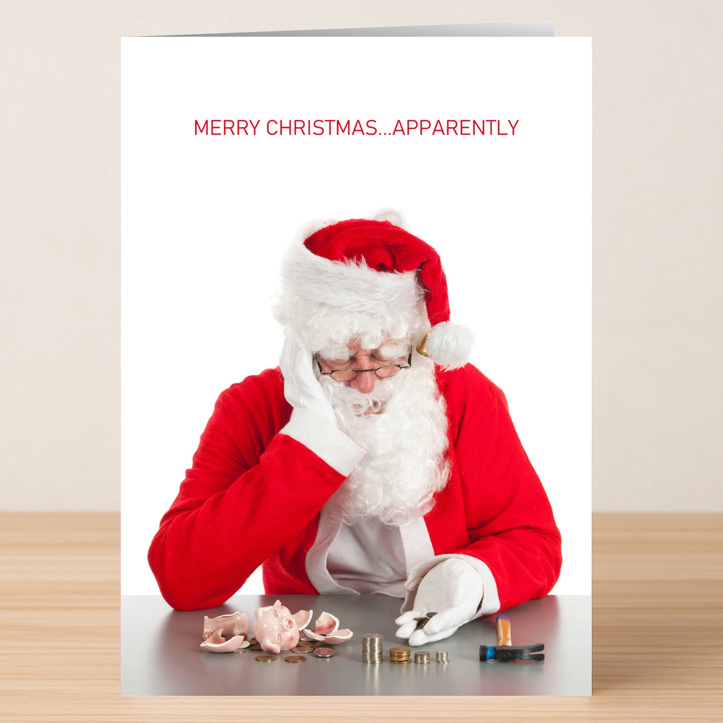 A person in a Santa costume sits at a table with coins and a broken piggy bank. The text above reads, "MERRY CHRISTMAS...APPARENTLY." Send the unexpected with the Apparently Funny Christmas Card from Twisted Gifts!.