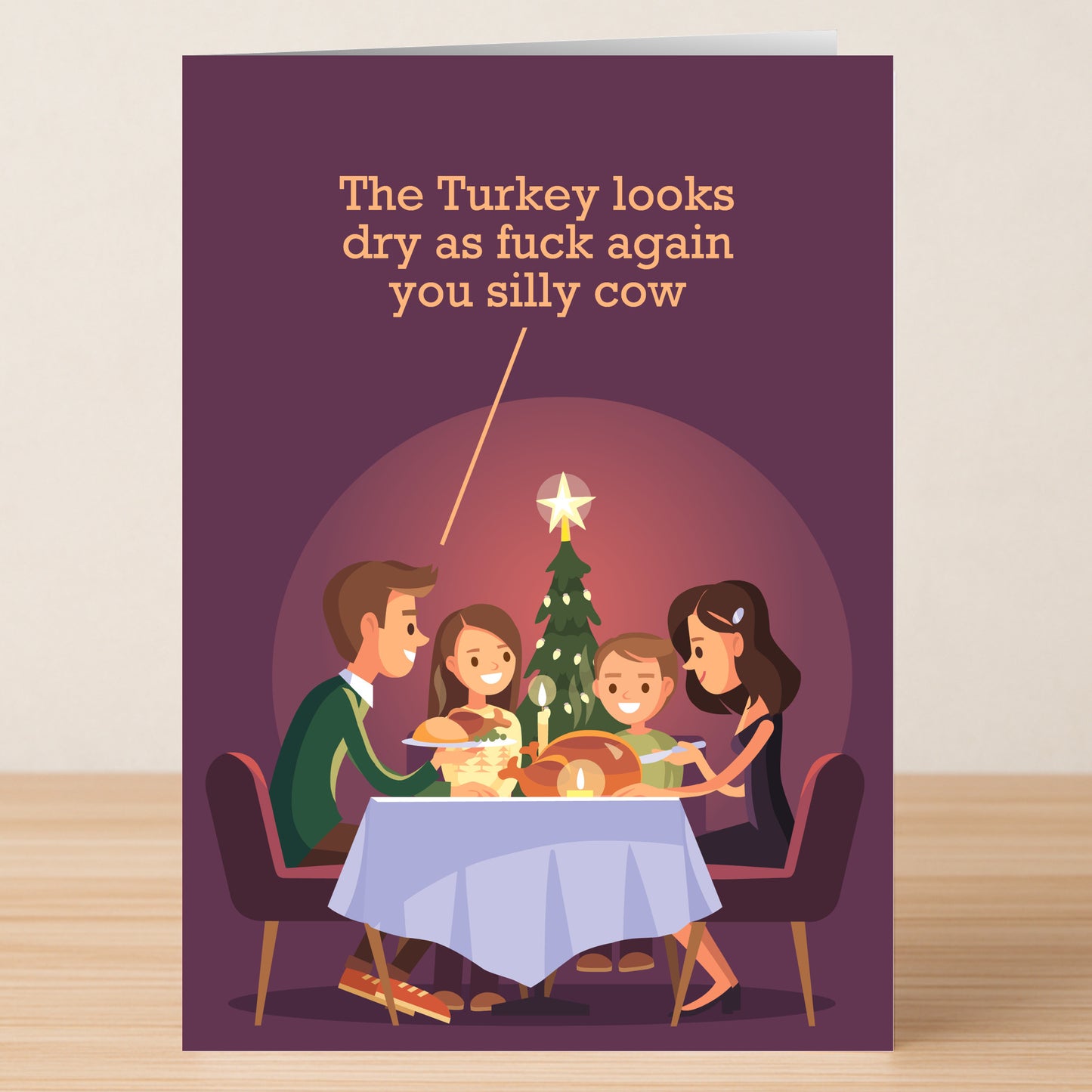 The Silly Cow Funny Christmas Card by Twisted Gifts features a cartoon family at the table with a Christmas tree, and the caption: "The Turkey looks dry as fuck again you silly cow." A perfect humorous card for sharing the holiday spirit.