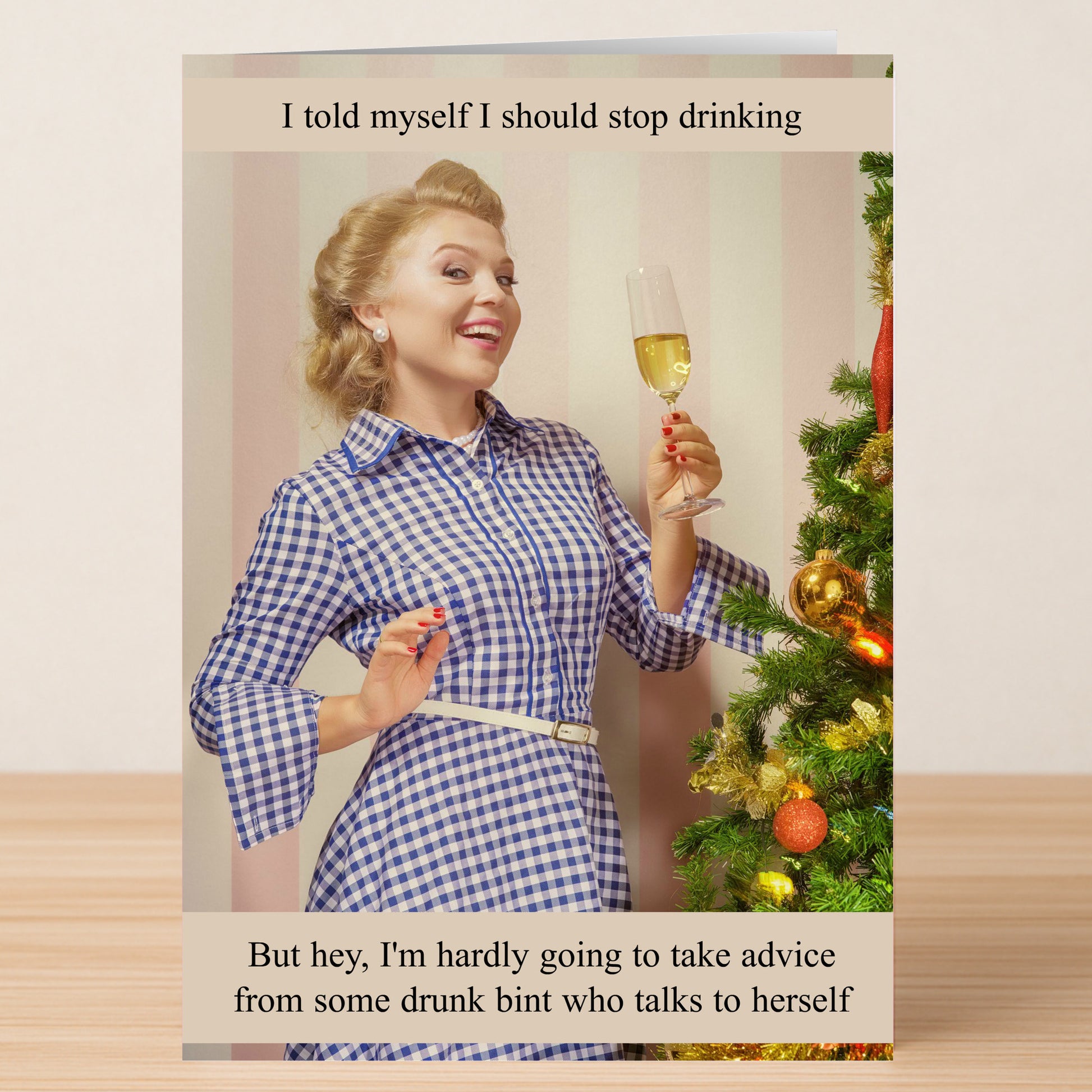 A woman in a blue checkered dress holding champagne smiles by a Christmas tree, with witty text above and below. The "Drunk Bint" Funny Christmas Card by Twisted Gifts perfectly captures festive humour for the holiday season.