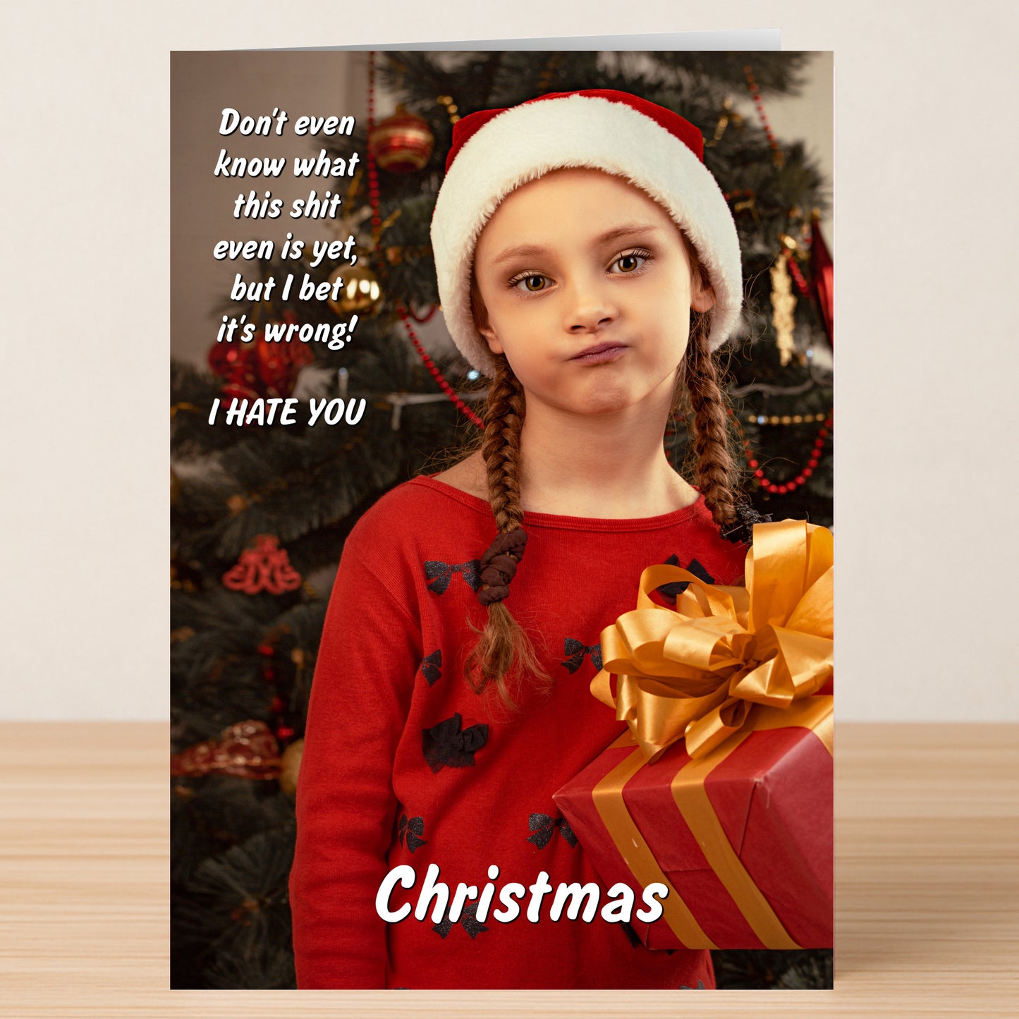 A young girl in a Santa hat and red dress holds a gift by the Christmas tree. This Twisted Gifts Christmas Card features a cheeky, humorous, and sarcastic holiday message—perfect for adding fun to your festivities.