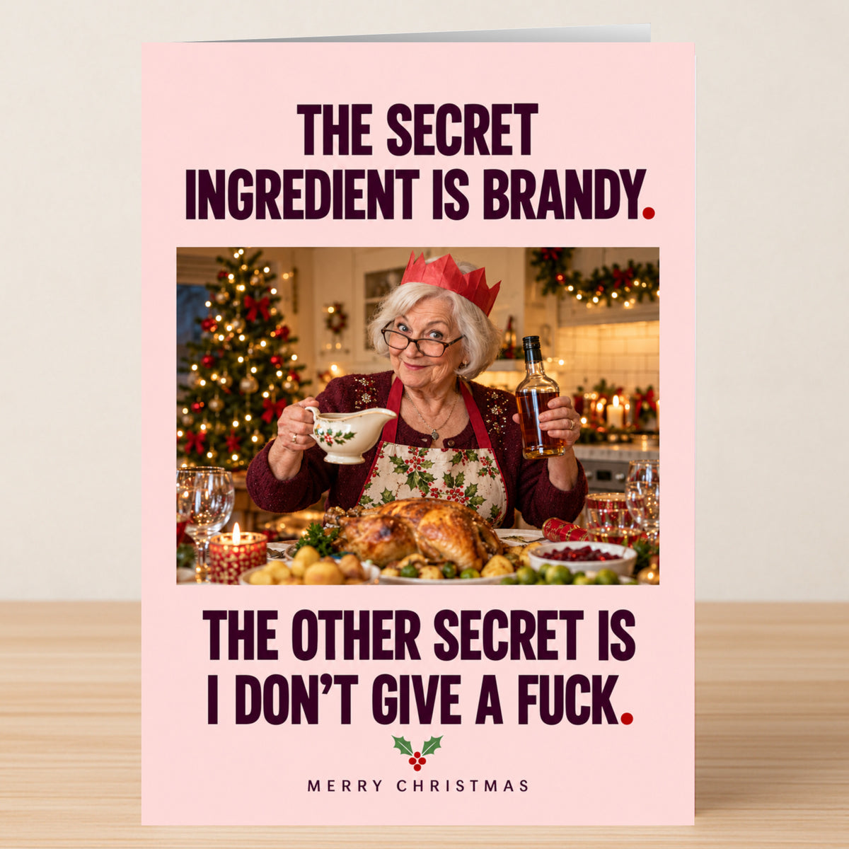 An older woman wearing a paper crown at a dinner table holds a gravy boat and brandy on this Secret Ingedient - Funny Christmas Card, which reads: “The secret ingredient is brandy. I don’t give a fuck.”