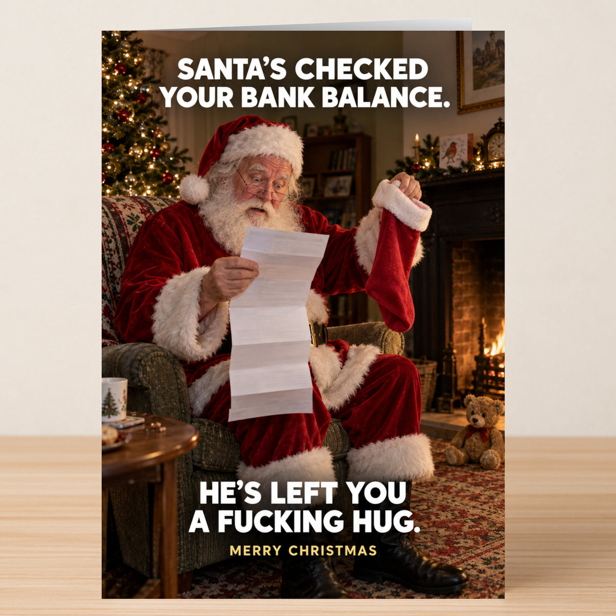 Bank Balance - Funny Christmas Card featuring Santa in an armchair by a fireplace holding a letter and a stocking, with text reading: “Santa’s checked your bank balance. He’s left you a fucking hug.”