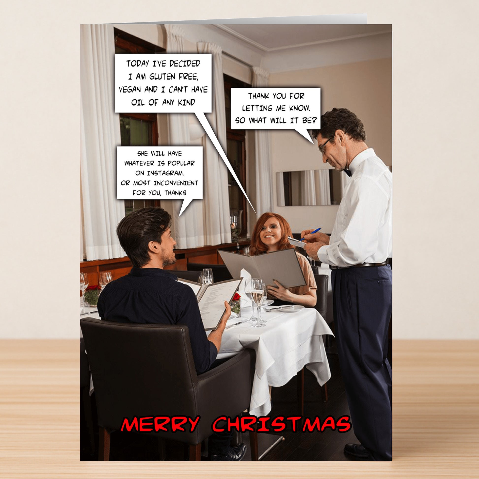 A couple at a restaurant shares preferences while ordering; the waiter listens with notepad in hand. Festive laughter rings out. "Merry Christmas" appears below, as featured on the Inconvenient Funny Christmas Card by Twisted Gifts.
