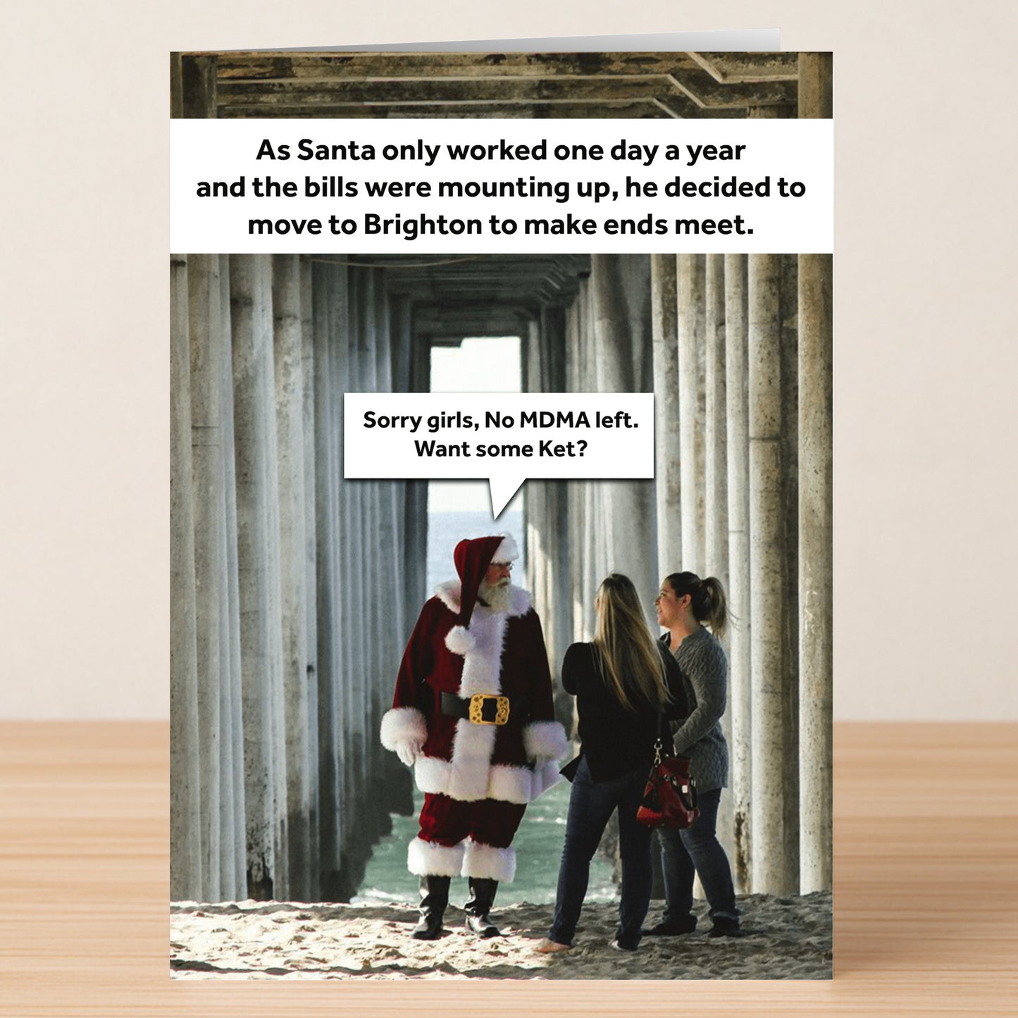 Santa Claus chats with two women under a pier in this Brighton Funny Christmas Card by Twisted Gifts, featuring witty captions about financial struggles and party drugs for a festive, humorous touch.