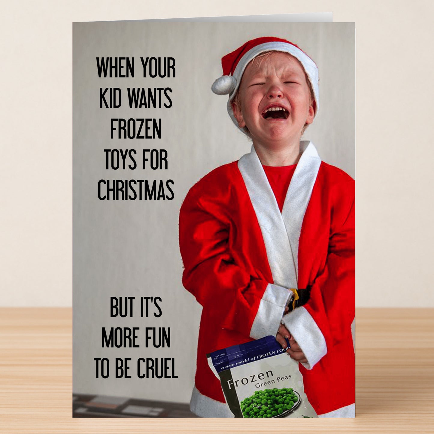 A child in a Santa costume cries while holding frozen peas. Text: "When your kid wants Frozen toys for Christmas, but it's more fun to be cruel." Frozen Funny Christmas Card by Twisted Gifts—perfect for your hilarious holiday greetings!.