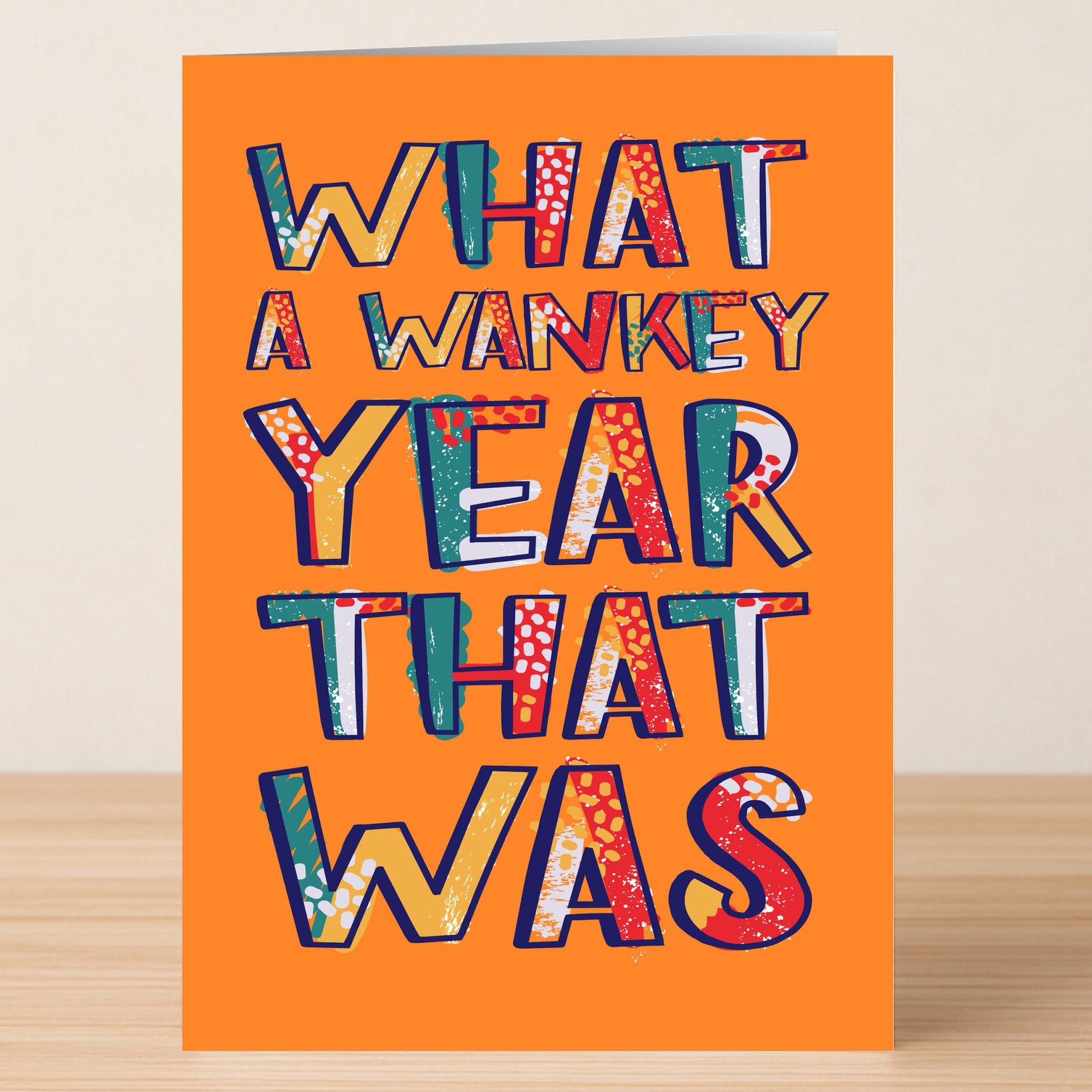 The Twisted Gifts "What A Year Funny Christmas Card" features bold, colorful text on orange card stock, adding humor to the holiday season as it stands on a wooden surface.