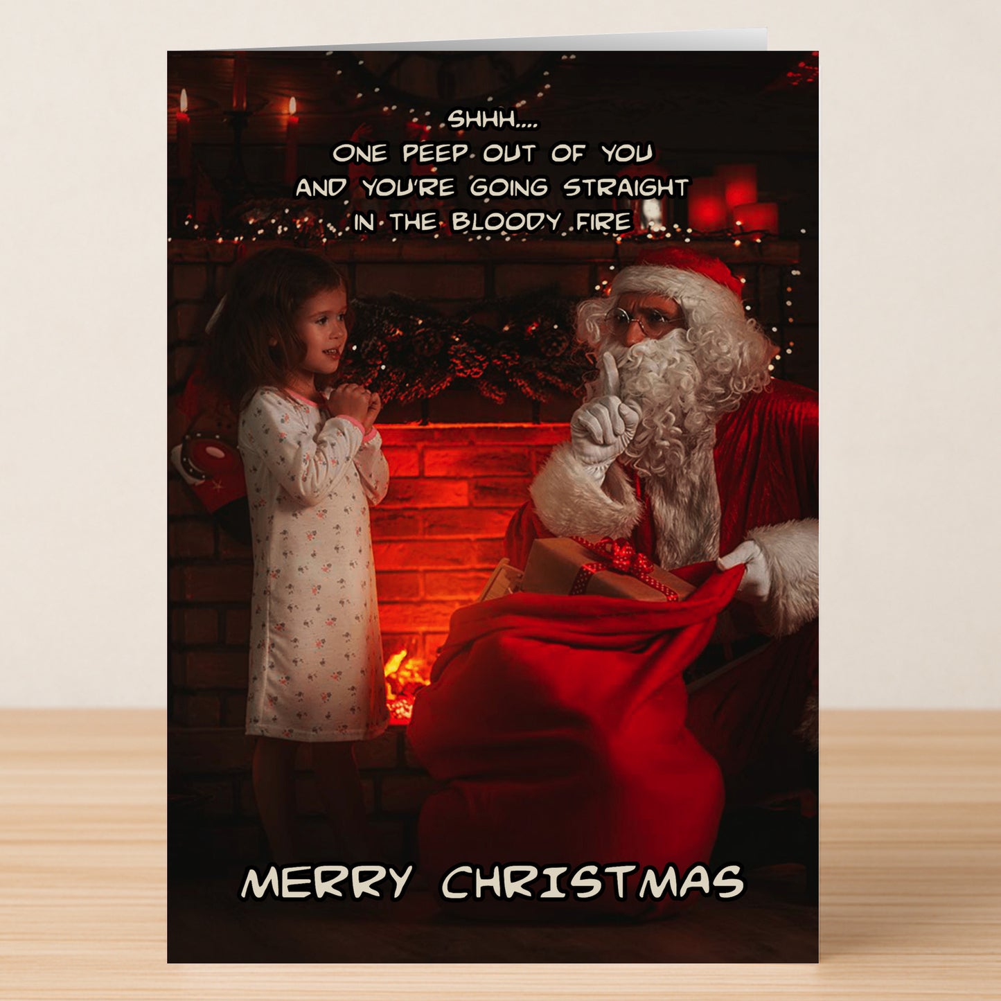 A young girl stands with Santa near a fireplace on the Twisted Gifts "Bloody Fire Funny Christmas Card," where Santa hushes her as a darkly funny message and "Merry Christmas" add twisted holiday cheer.