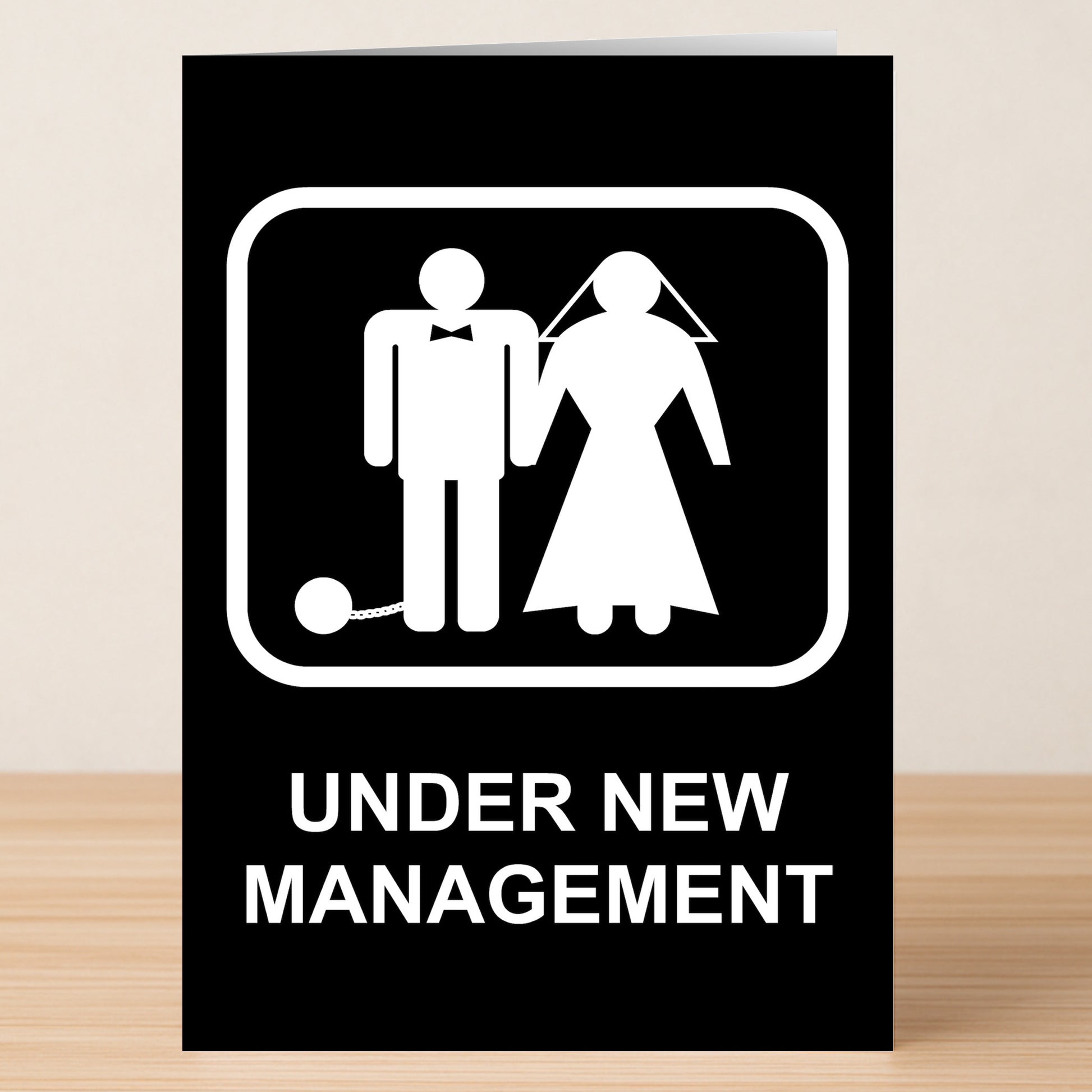 The Twisted Gifts Under New Management Funny Congratulations Card features bride and groom icons, the groom with a ball and chain, and "UNDER NEW MANAGEMENT" text—an ideal humorous card for congratulating newlyweds.