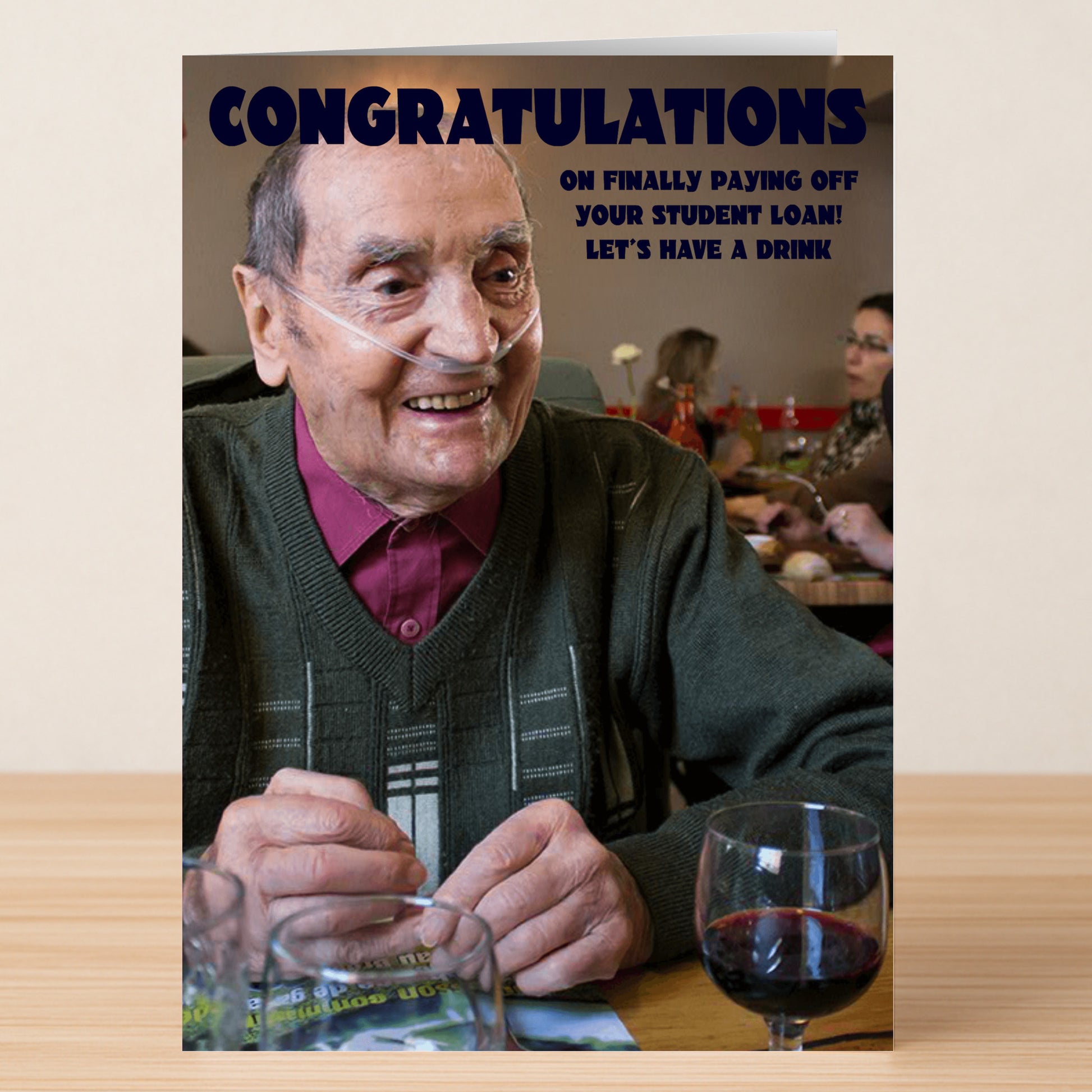 An elderly man with oxygen tubing smiles at a table with wine and a Twisted Gifts "Student Loan Funny Congratulations Card" that reads, "Congratulations on finally paying off your student loan! Let's celebrate accomplishment with a drink.