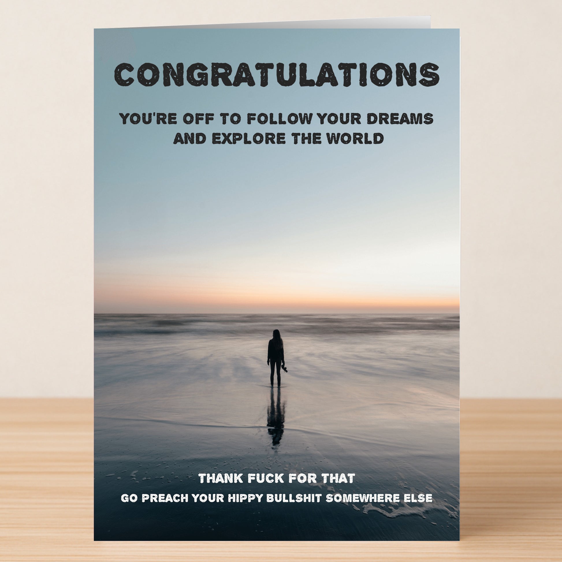 The "Explore The World Insulting Congratulations Card" by Twisted Gifts features a silhouetted beach walker at sunset and reads, "THANK FUCK FOR THAT. GO PREACH YOUR HIPPY BULLSHIT SOMEWHERE ELSE.