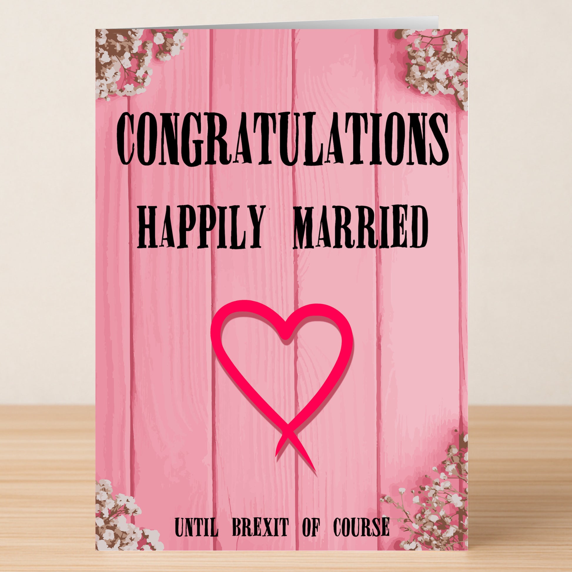The Happily Married Funny Congratulations Card by Twisted Gifts features a pink design with "CONGRATULATIONS HAPPILY MARRIED" and "UNTIL BREXIT OF COURSE," plus heart and floral accents on a wooden background.