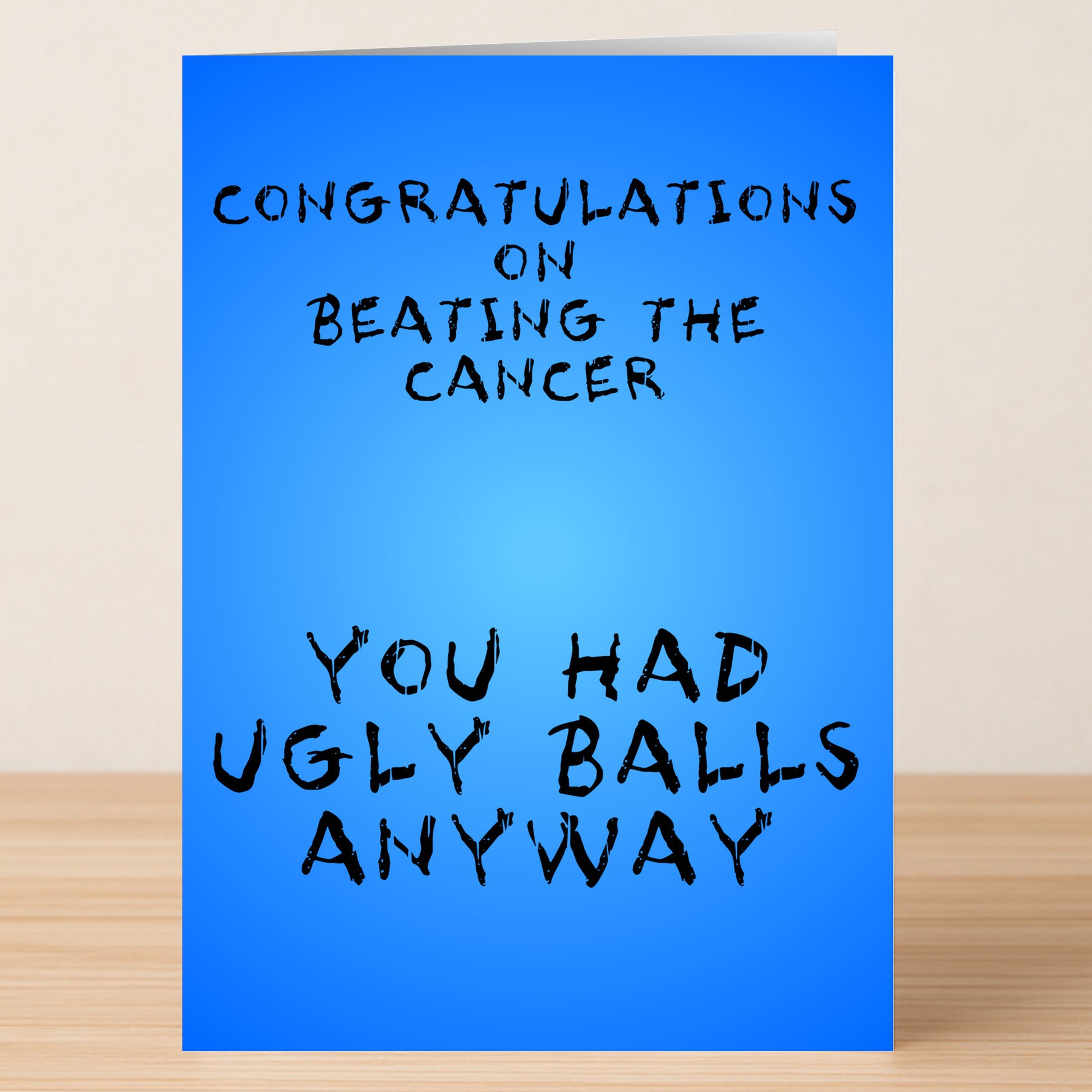 The Ugly Balls Insulting Congratulations Card by Twisted Gifts features a blue design that reads, "Congratulations on beating the cancer. You had ugly balls anyway," displayed on a wooden surface.