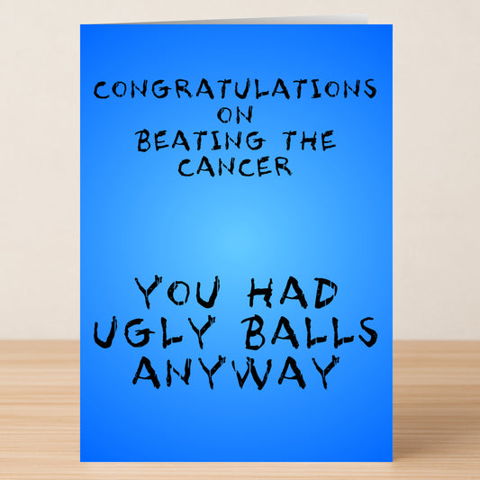The Ugly Balls Insulting Congratulations Card by Twisted Gifts features a blue design that reads, "Congratulations on beating the cancer. You had ugly balls anyway," displayed on a wooden surface.