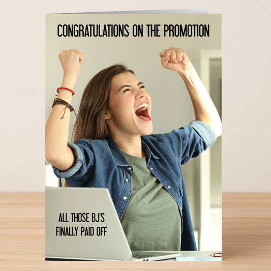 A woman celebrates at her desk with a laptop on the Paid Off Rude Congratulations Card by Twisted Gifts, featuring the cheeky message: "Congratulations on the promotion. All those BJ's finally paid off.