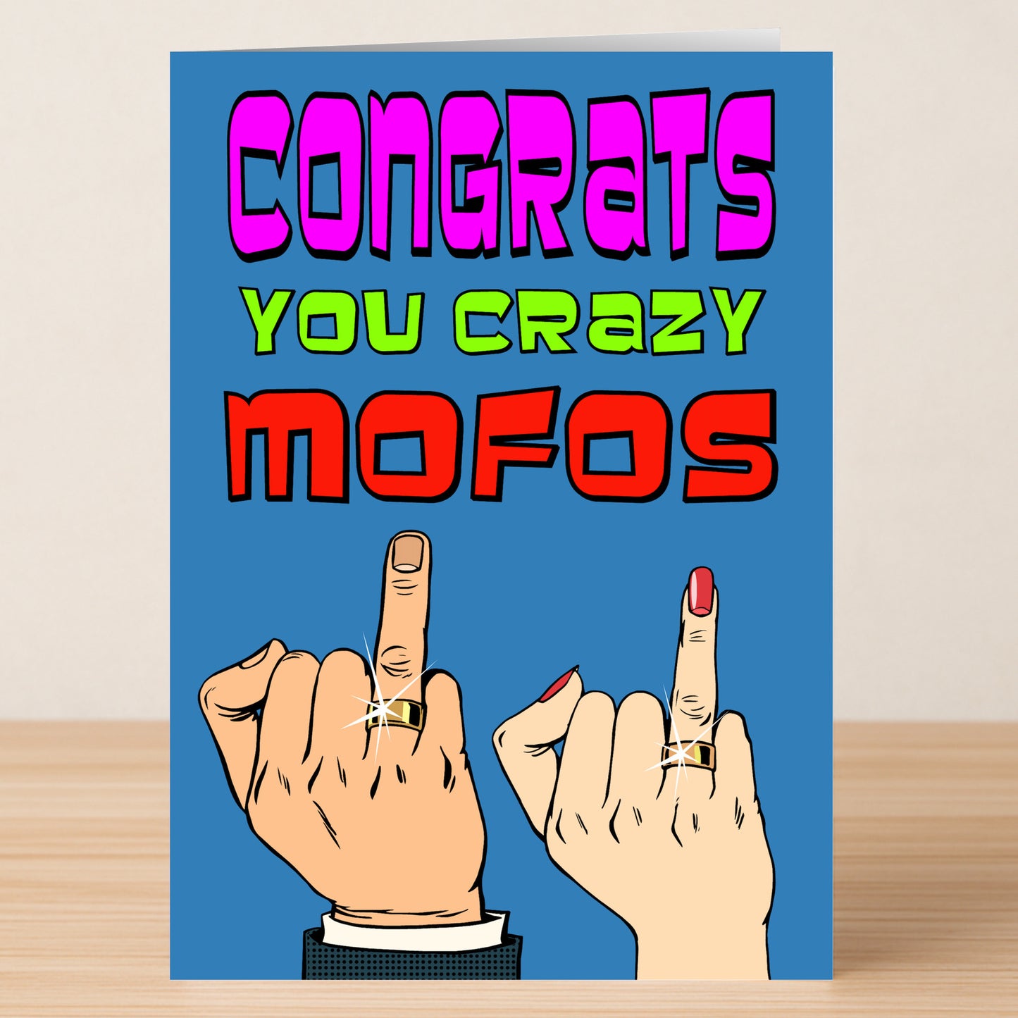 The Mofos Insulting Congratulations Card by Twisted Gifts features bold "Congrats You Crazy Mofos" text, two hands flipping the bird with rings, and a blue background—ideal for friends who love explicit language and cheeky humor.
