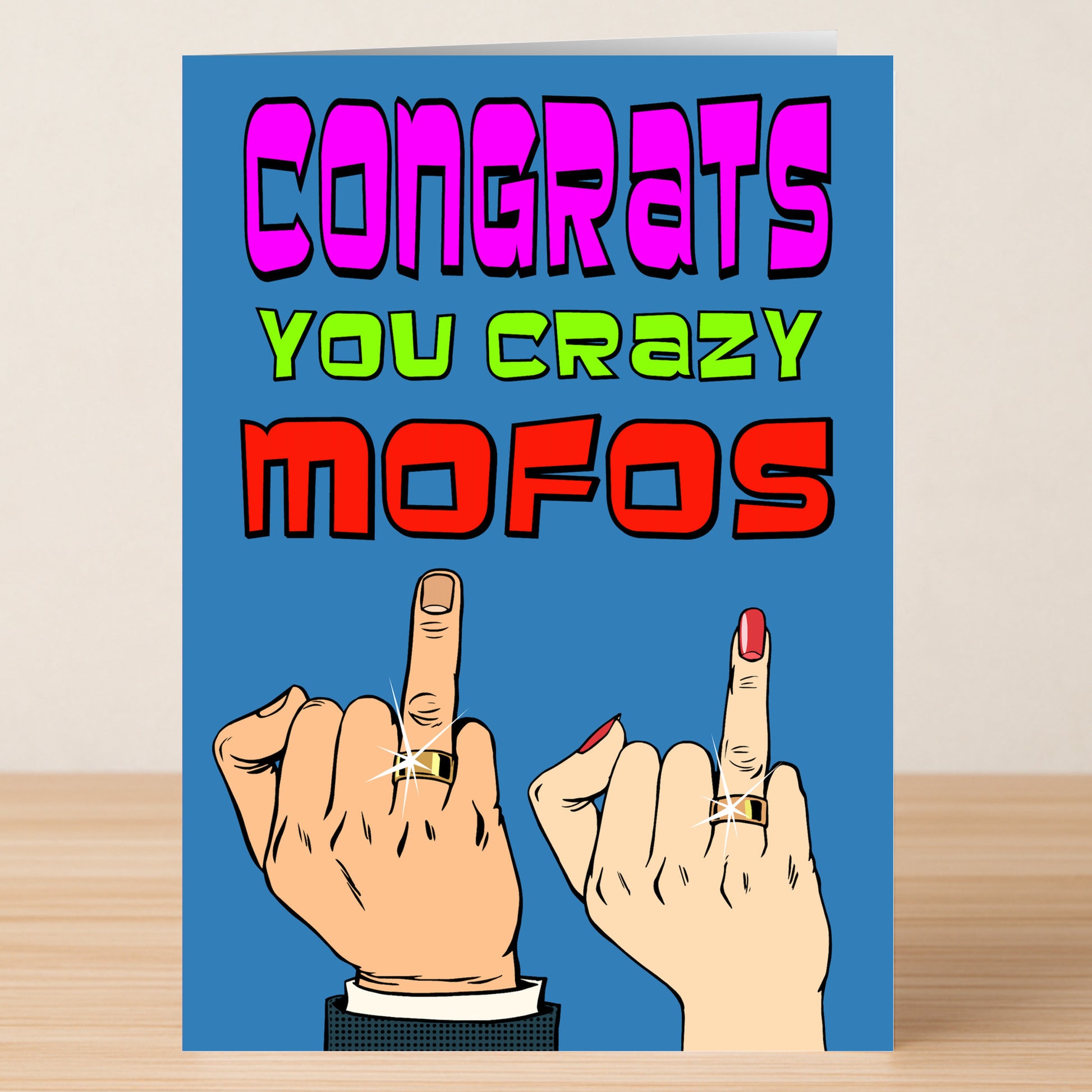 The Mofos Insulting Congratulations Card by Twisted Gifts features bold "Congrats You Crazy Mofos" text, two hands flipping the bird with rings, and a blue background—ideal for friends who love explicit language and cheeky humor.