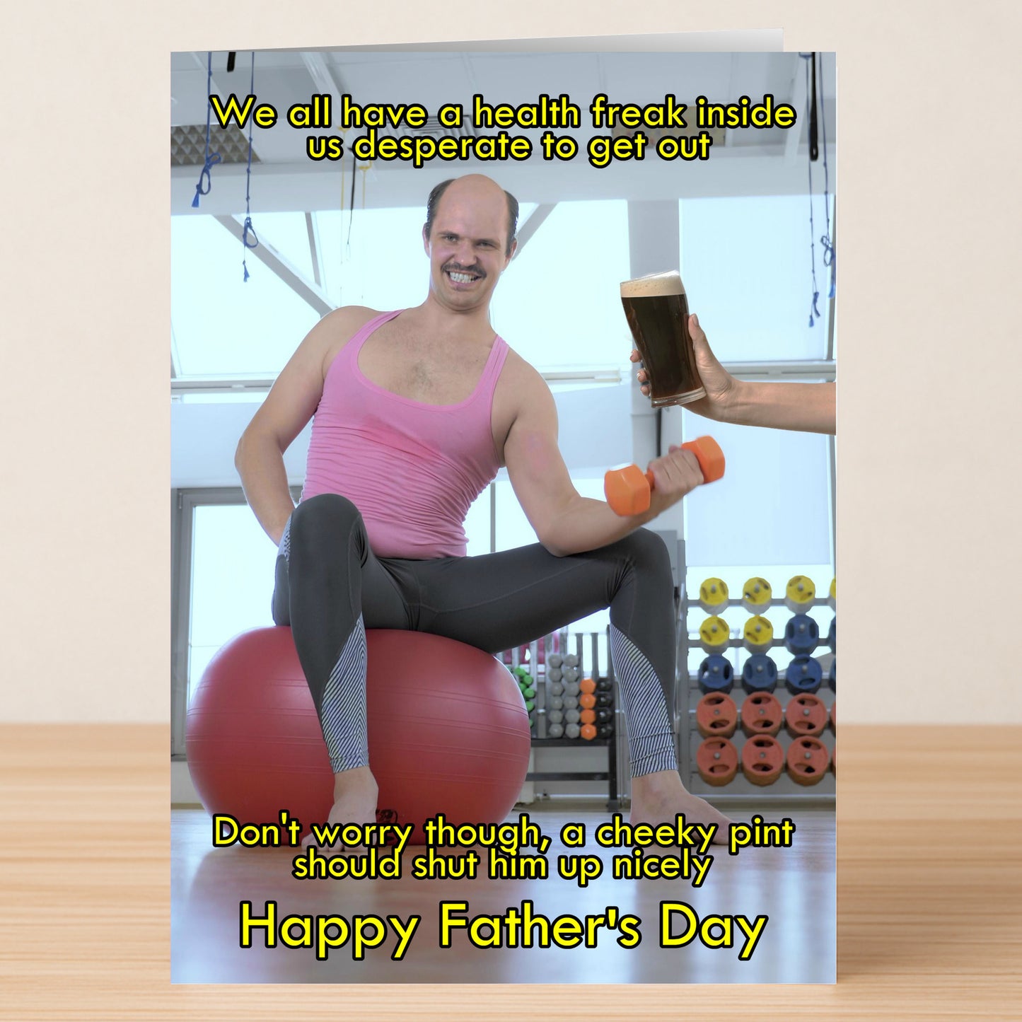 A man in workout gear smiles while lifting a dumbbell on an exercise ball as a beer is handed to him—the Cheeky Pint Funny Day Card by Twisted Gifts, perfect for Father’s Day with its humorous touch.