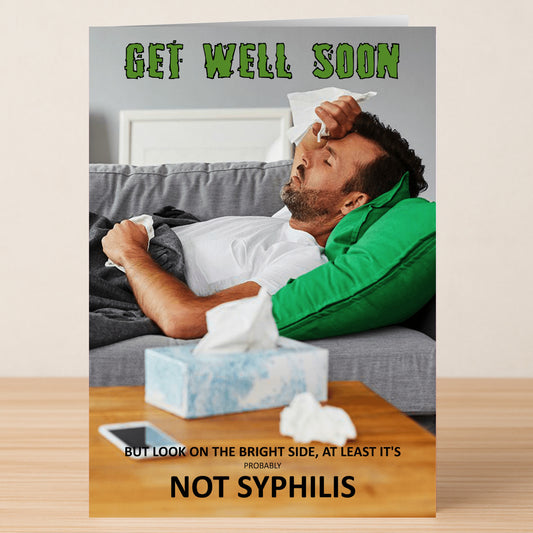 A man lies on a couch looking unwell, tissues nearby. Text: "Get well soon. But look on the bright side, at least it's probably not syphilis." The Syphilis Rude Get Well Soon Card by Twisted Gifts adds humor to recovery.