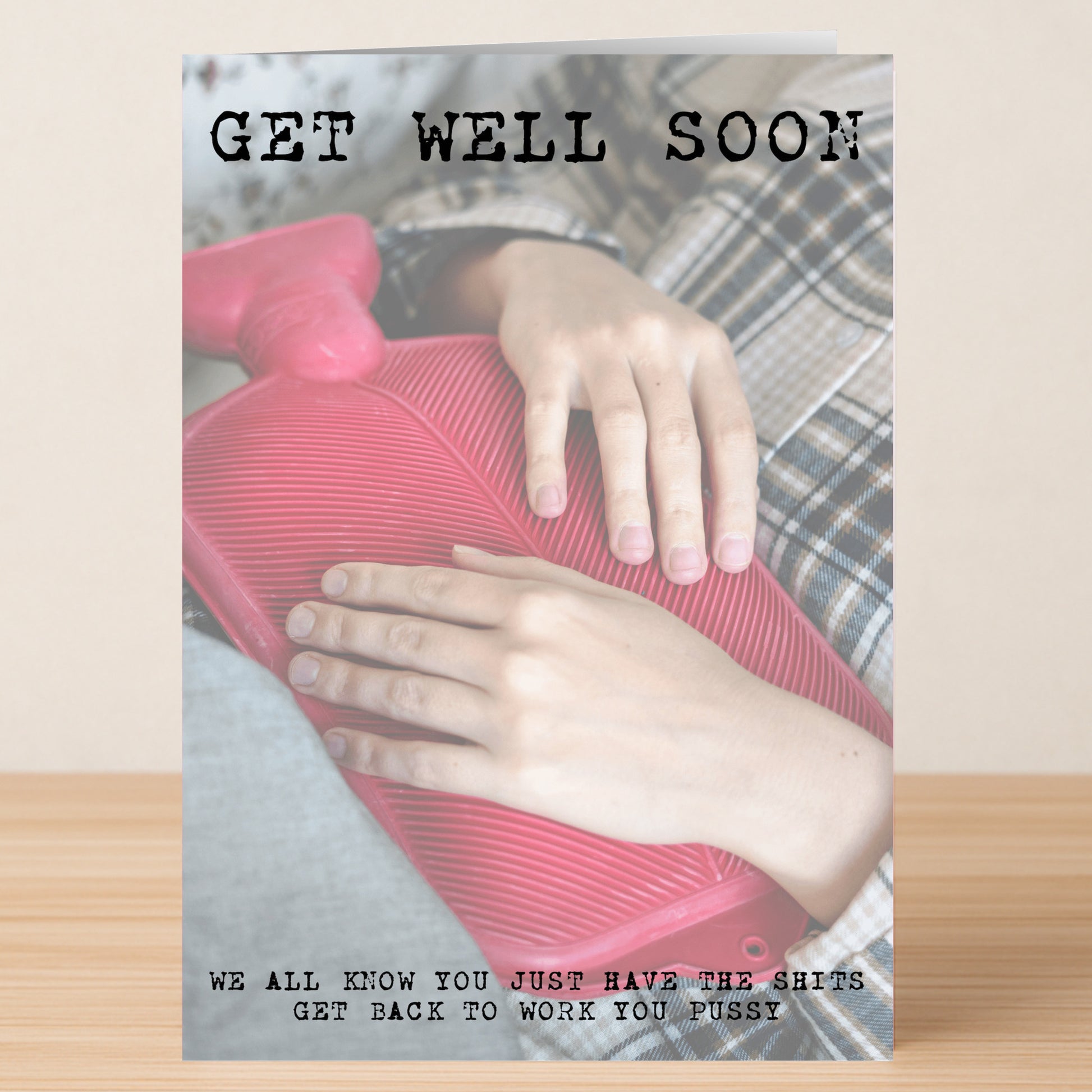 A person in plaid pajamas holds a red hot water bottle with “The Shits Insulting Get Well Soon Card” by Twisted Gifts, featuring a funny message about getting back to work.