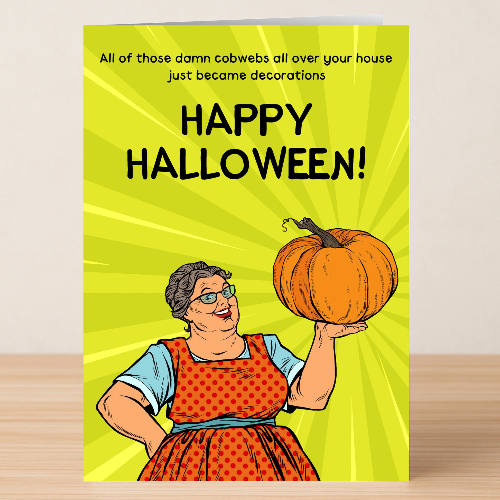 A woman in glasses and a red apron holds a pumpkin as the Twisted Gifts Cobwebs Insulting Halloween Card reads, “All those damn cobwebs all over your house just became decorations. Happy Halloween!”—perfect for your spooky celebrations!.