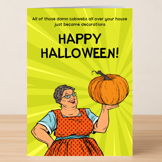 A woman in glasses and a red apron holds a pumpkin as the Twisted Gifts Cobwebs Insulting Halloween Card reads, “All those damn cobwebs all over your house just became decorations. Happy Halloween!”—perfect for your spooky celebrations!.