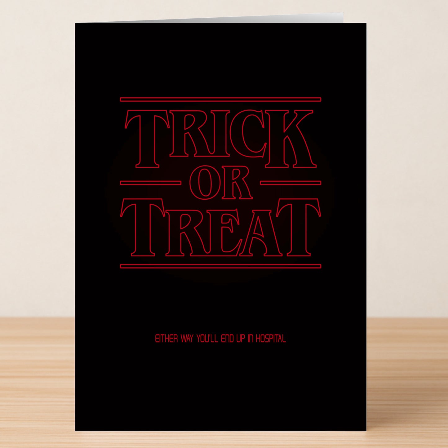 The Stranger Funny Halloween Card by Twisted Gifts features bold red "Trick or Treat" text, smaller "Either way you'll end up in hospital," a hilarious design, and is pictured standing on a wooden surface.
