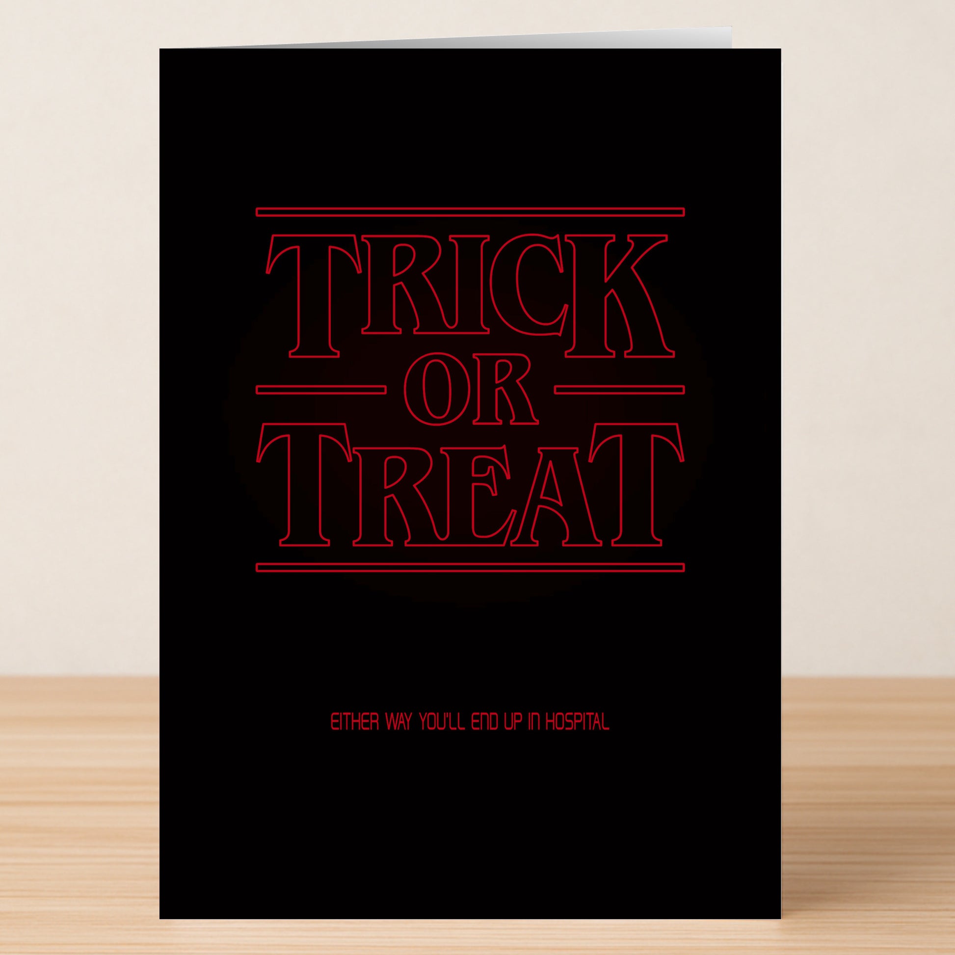 The Stranger Funny Halloween Card by Twisted Gifts features bold red "Trick or Treat" text, smaller "Either way you'll end up in hospital," a hilarious design, and is pictured standing on a wooden surface.