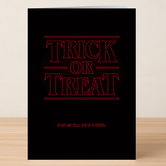 The Stranger Funny Halloween Card by Twisted Gifts features bold red "Trick or Treat" text, smaller "Either way you'll end up in hospital," a hilarious design, and is pictured standing on a wooden surface.