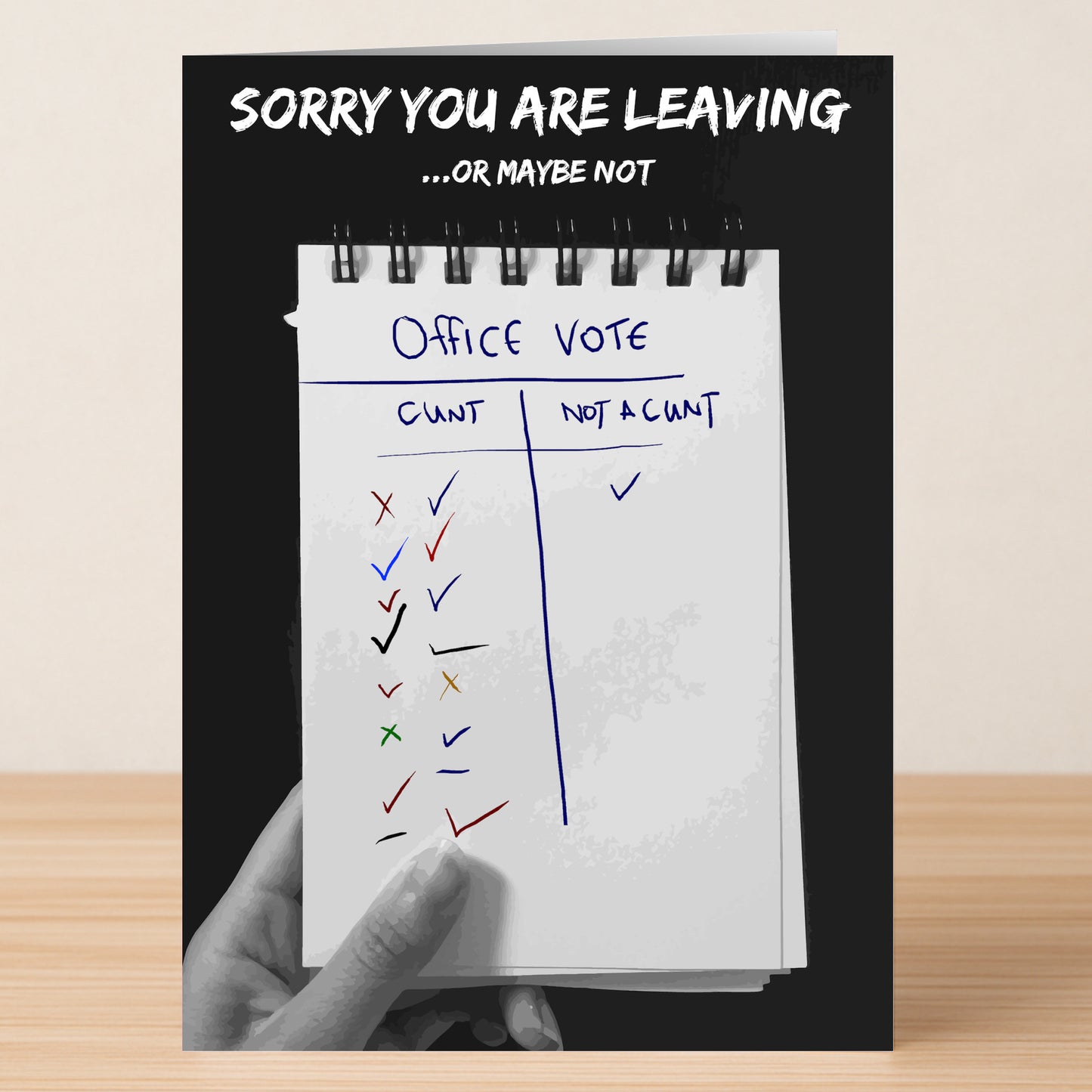 A hand holds Twisted Gifts' "Office Vote – Insulting Sorry Card," a notepad-styled farewell with columns "CUNT" and "NOT A CUNT," and the message, “SORRY YOU ARE LEAVING ...OR MAYBE NOT”—ideal for those with rude humour.