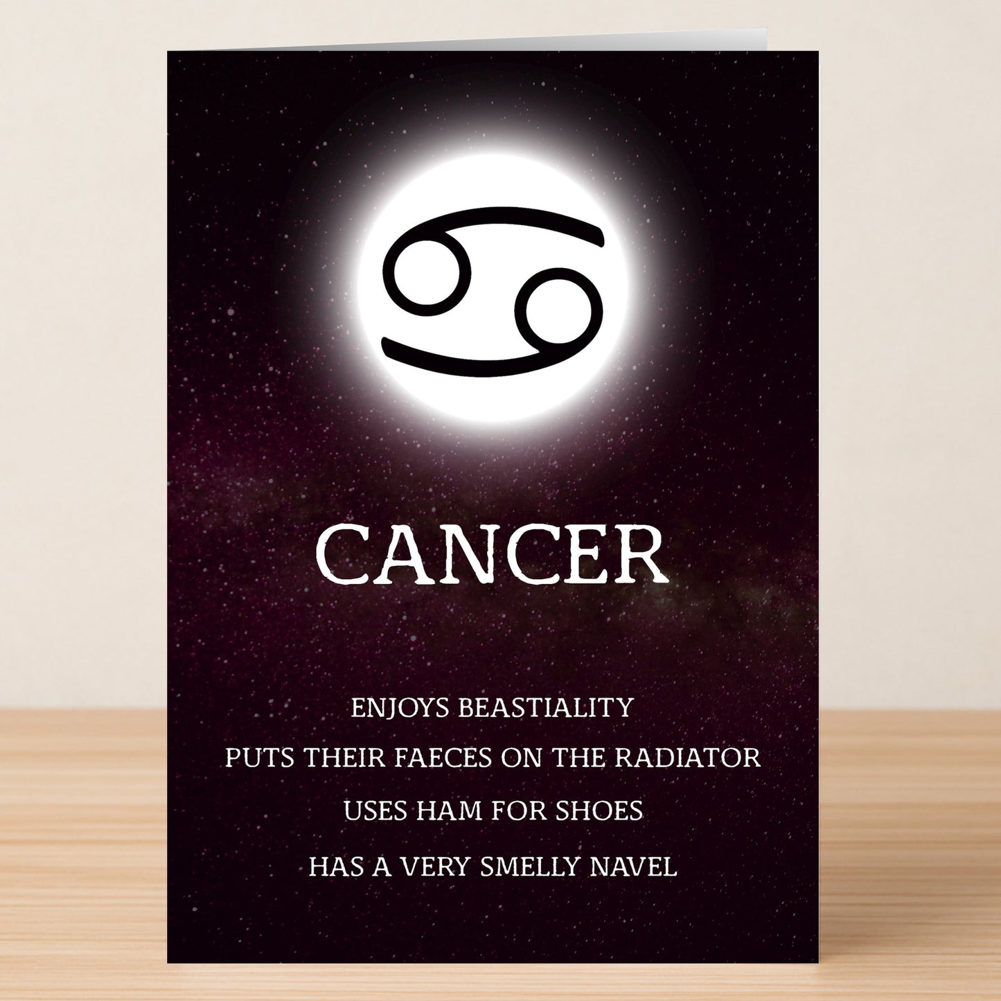 The Cancer – Rude Zodiac Greeting Card by Twisted Gifts features the Cancer symbol and name with outrageous, funny traits on a dark starry background—a bold astrology card shown standing on a wooden surface.