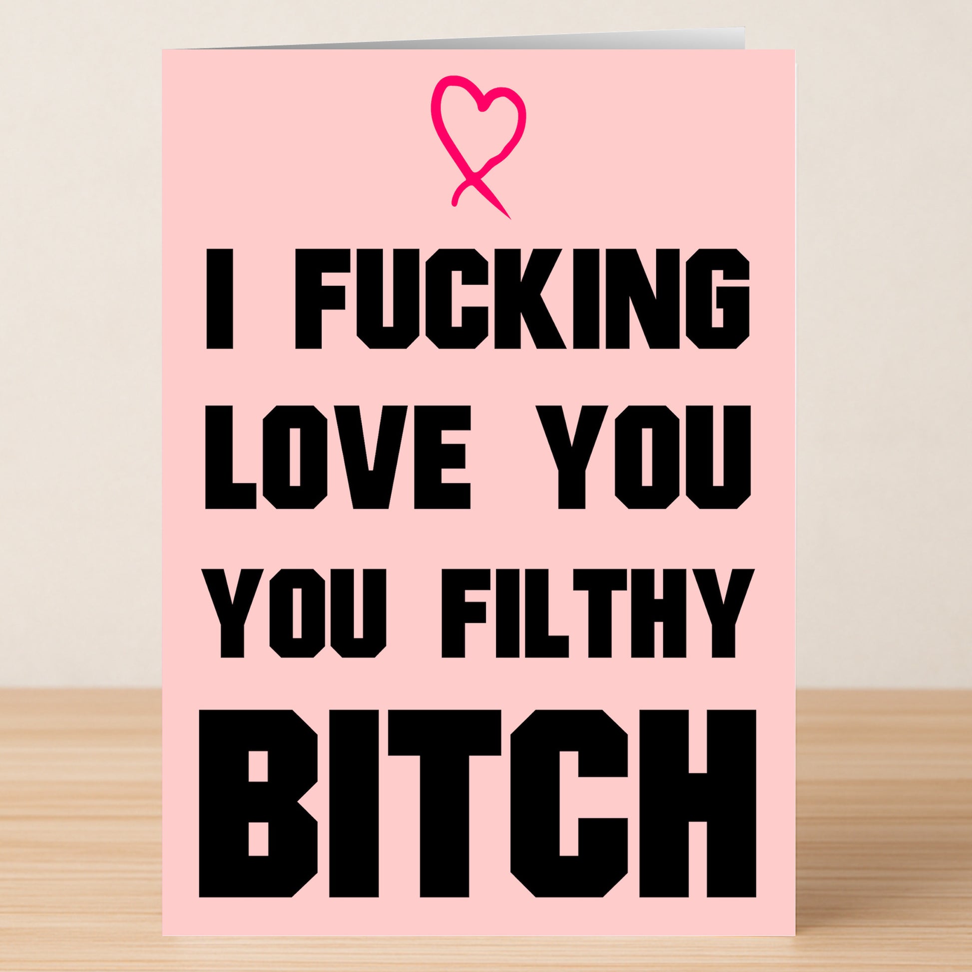 The Filthy Bitch Rude Greeting Card by Twisted Gifts is a pink card with a heart and bold black text saying, "I fucking love you you Filthy Bitch," shown on a wooden surface.