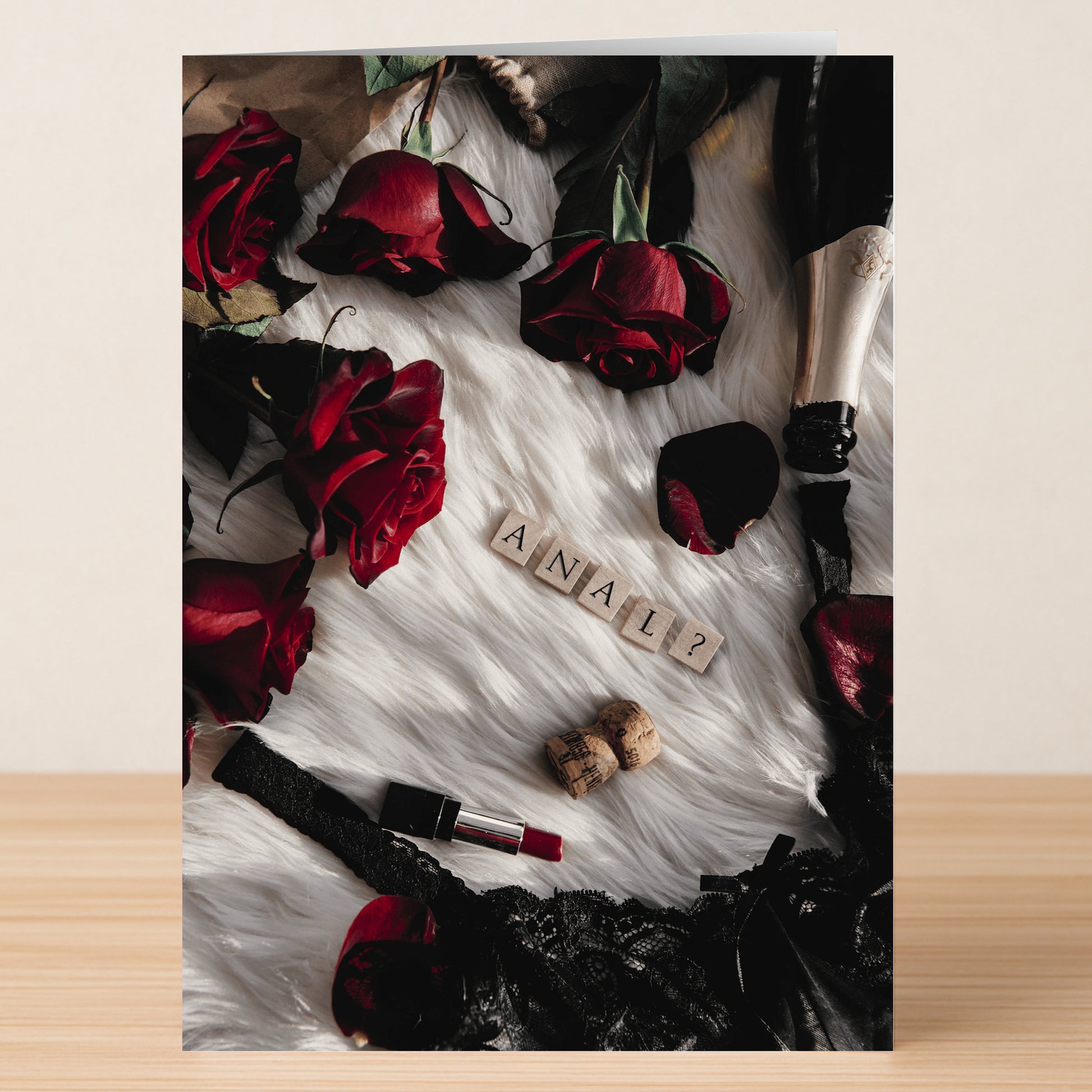 The Champagne and Roses Rude Valentine's Card by Twisted Gifts, set on a wooden surface with red roses, black lace lingerie, champagne, and Scrabble tiles spelling "ANAL?", adds humor and love—perfect for a cheeky celebration.