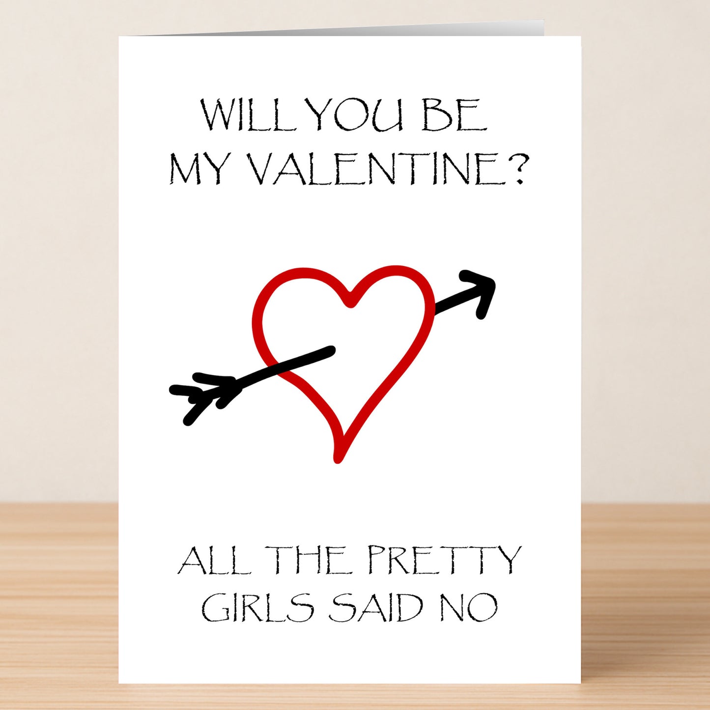 Pretty Insulting Valentine's Card by Twisted Gifts: red heart pierced by a black arrow, with the witty note, "Will you be my valentine? All the pretty girls said no." The card is displayed on a wooden surface.