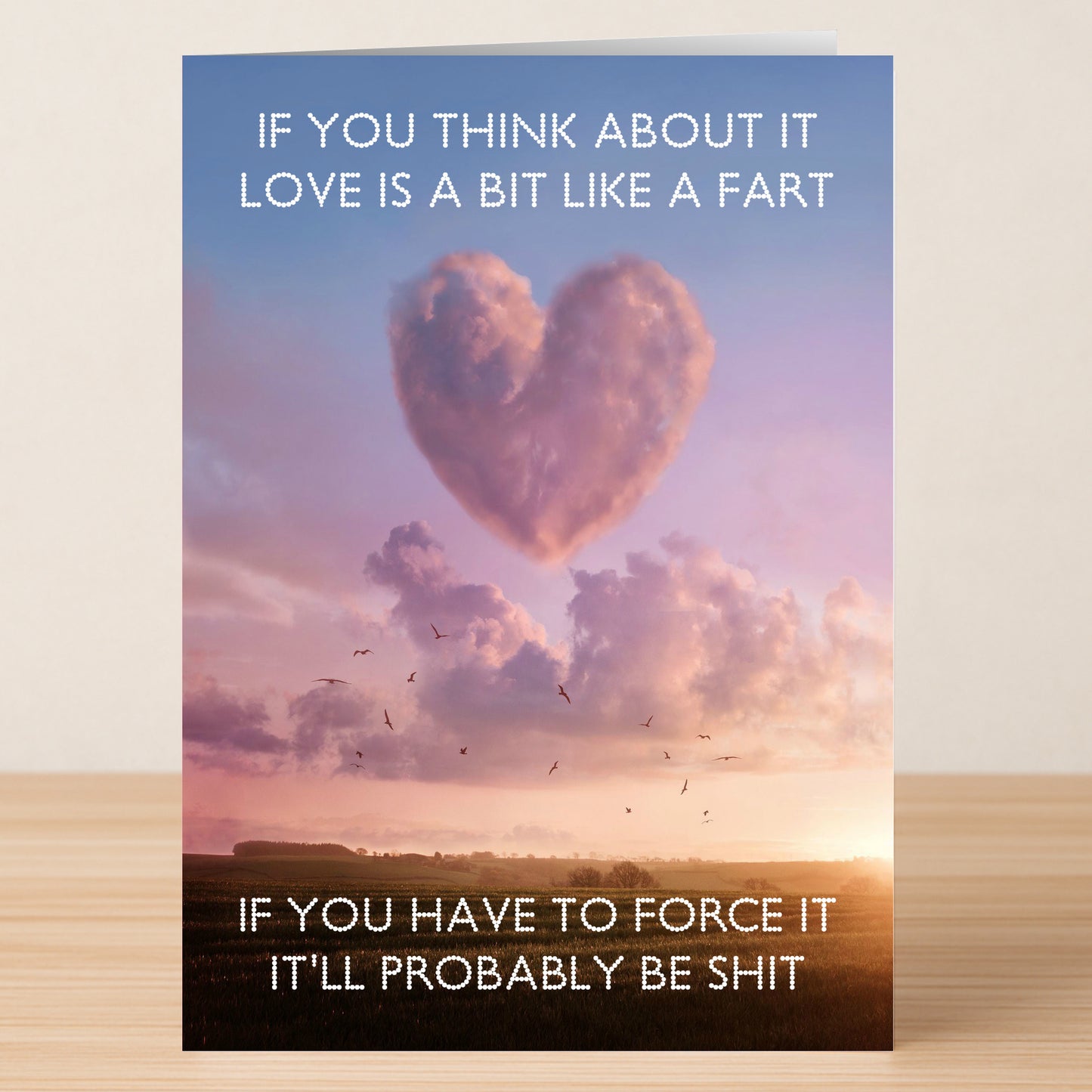 A Fart Funny Valentines Card by Twisted Gifts—features a heart-shaped cloud at sunset and playful text comparing love to a fart: laugh, don't force it, or it'll lead to trouble!.