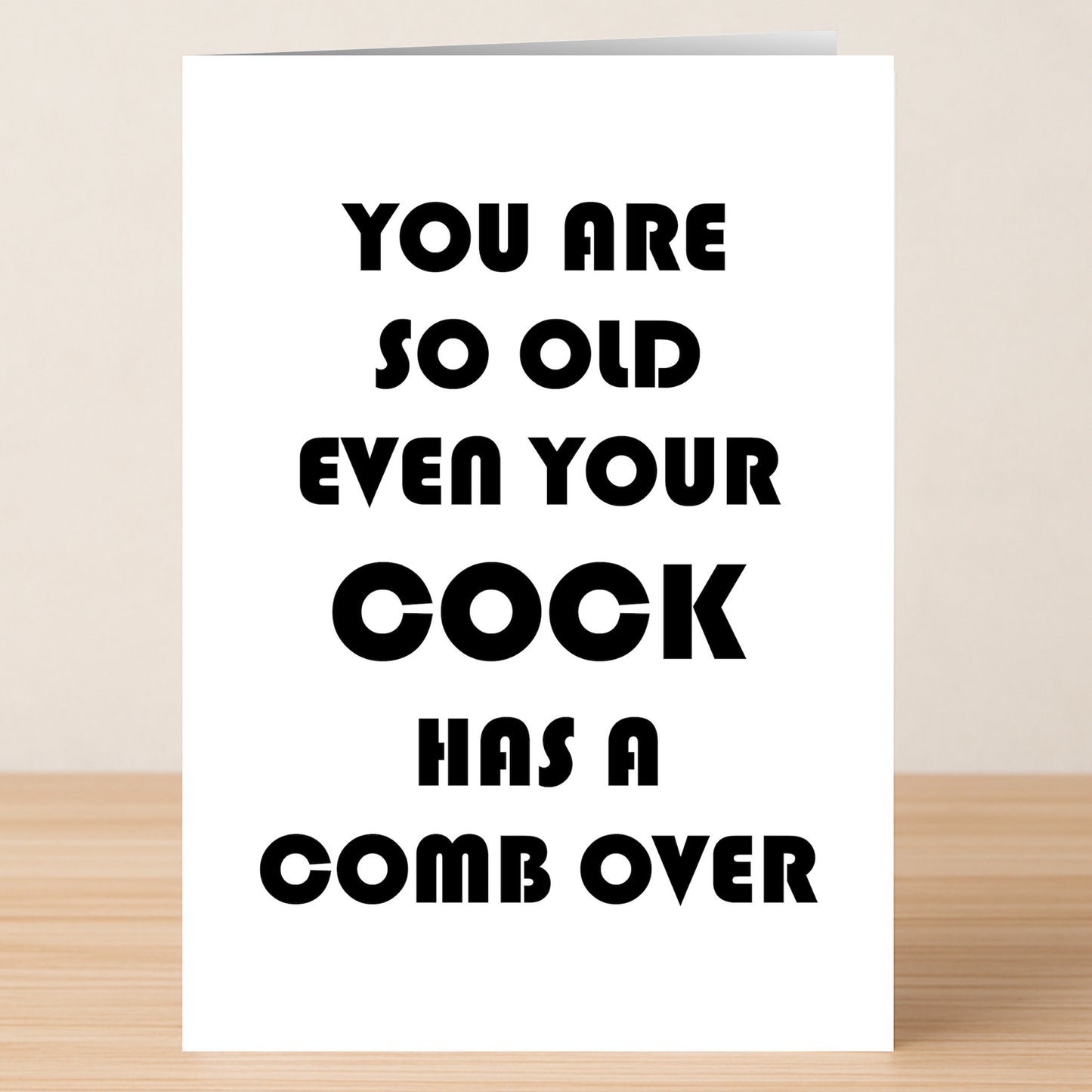 The "Comb Over – Insulting Birthday Card" from Twisted Gifts sits on a wooden surface, reading: "You are so old even your cock has a comb over." Ideal for anyone who enjoys wickedly funny birthday cards.