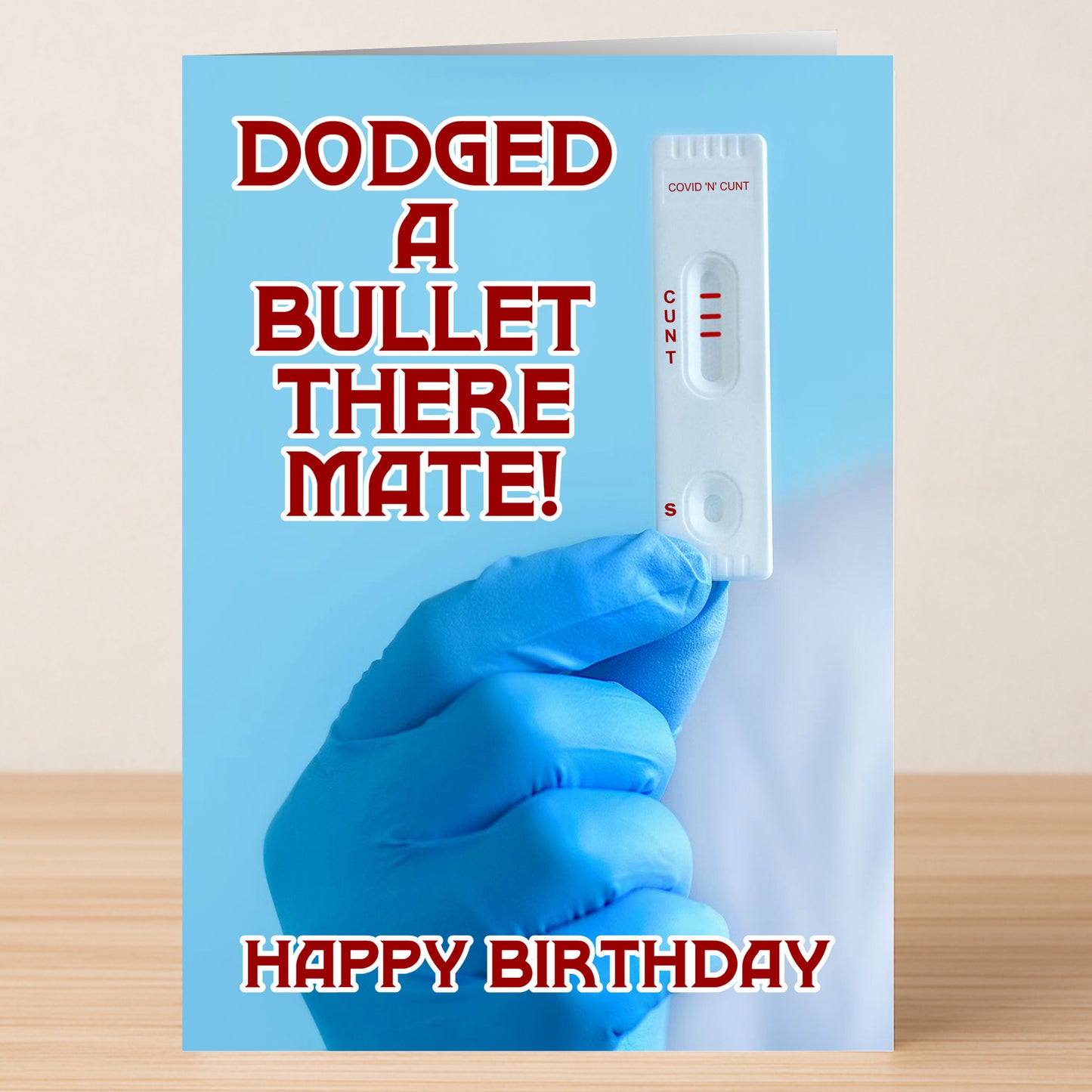 A gloved hand holds a COVID-19 test showing a negative result. Text reads, "Dodged a bullet there mate! Happy Birthday." The Dodged a Bullet – Insulting Birthday Card by Twisted Gifts is perfect for anyone who loves cheeky humor.