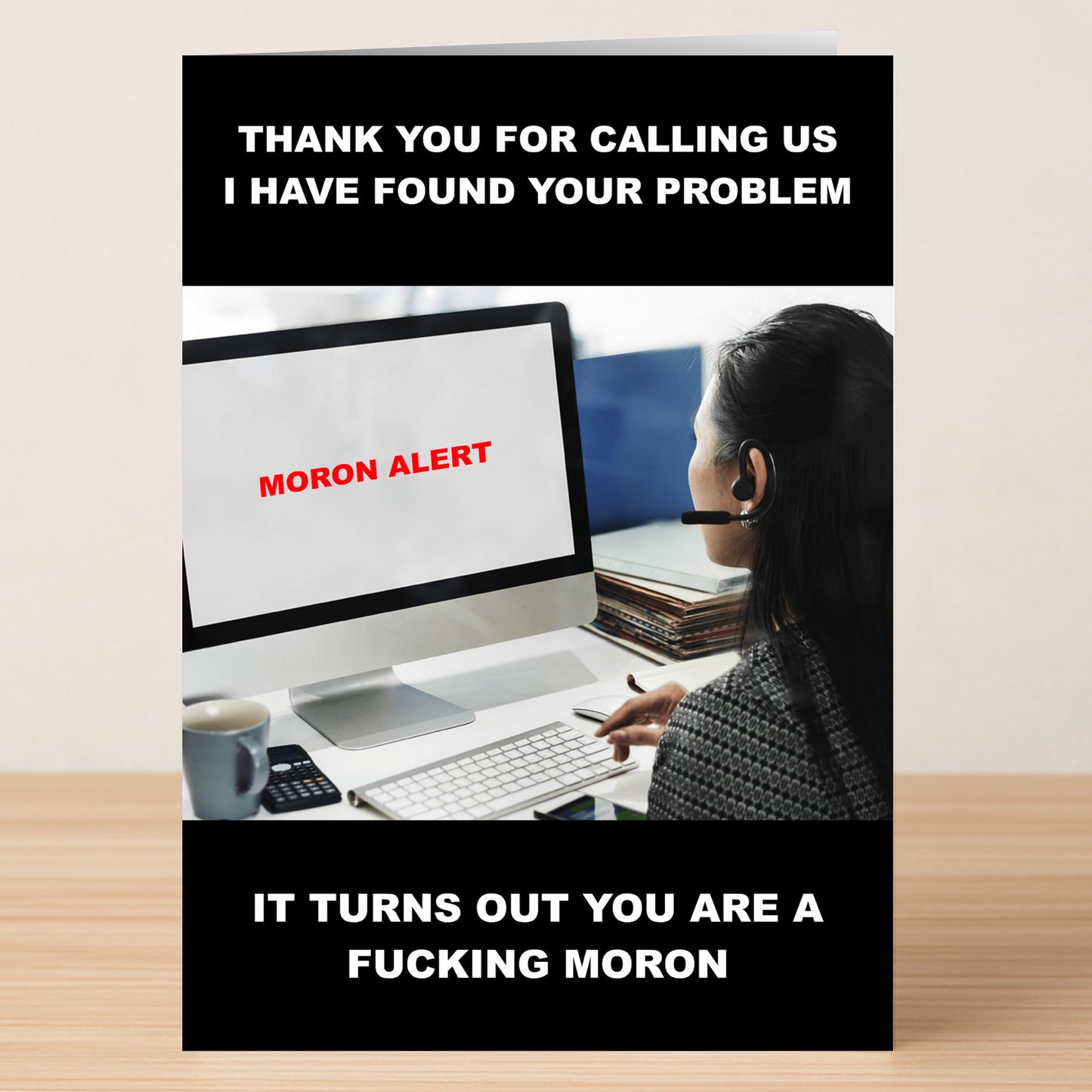A woman in a headset sits at her desk, looking at a screen showing "MORON ALERT"—a funny scene featured on the Moron Alert Insulting Greeting Card by Twisted Gifts, ideal for anyone who enjoys sarcastic and witty messages.